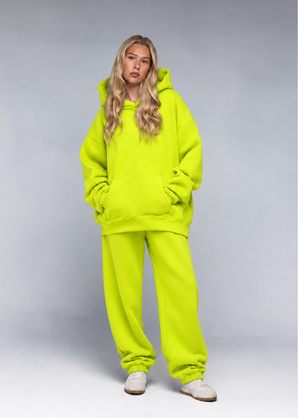 A woman modeling a matching neon yellow-green oversized hoodie and sweatpants set, paired with white sneakers, against a neutral gray studio background.