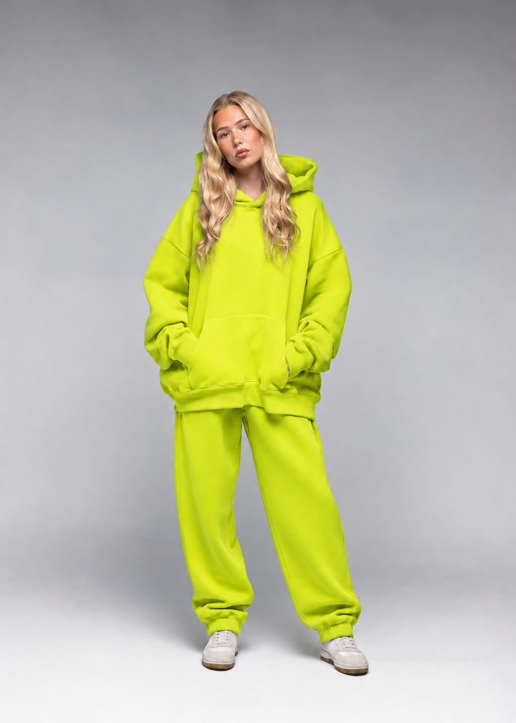 A woman modeling a matching neon yellow-green hooded sweatshirt and sweatpants set against a neutral gray studio background.