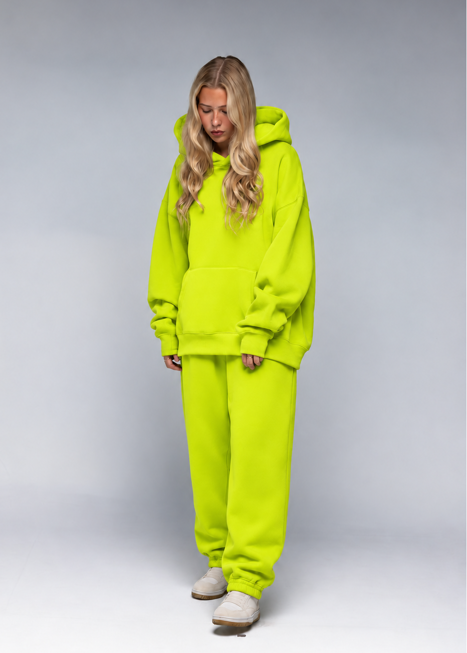 A woman wearing a bright neon yellow-green matching lounge set consisting of an oversized pullover hoodie and matching sweatpants, standing against a neutral gray studio background.