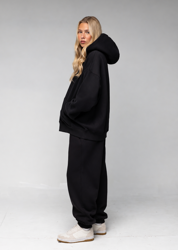 A woman modeling a black oversized hoodie and matching black sweatpants with white sneakers, standing against a light gray studio background.
