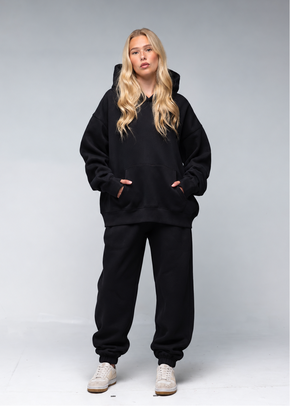 A blonde woman modeling a black oversized hoodie and matching black sweatpants against a light gray studio background.