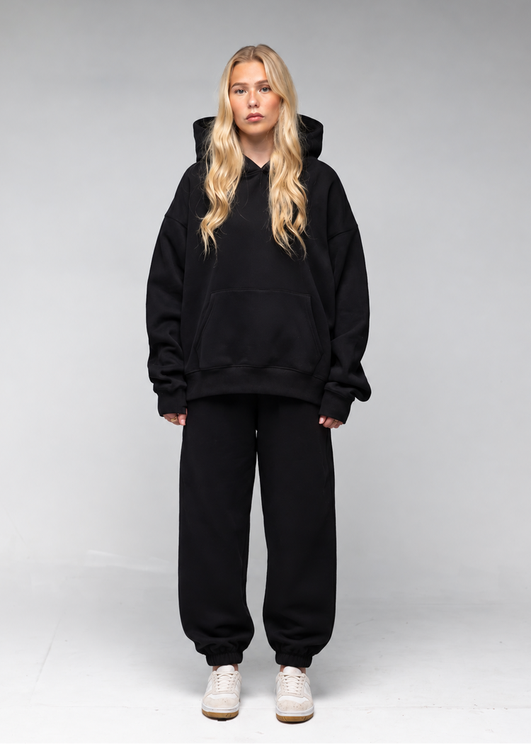A blonde woman stands against a light gray studio background wearing an oversized black hoodie, matching black sweatpants, and white sneakers.