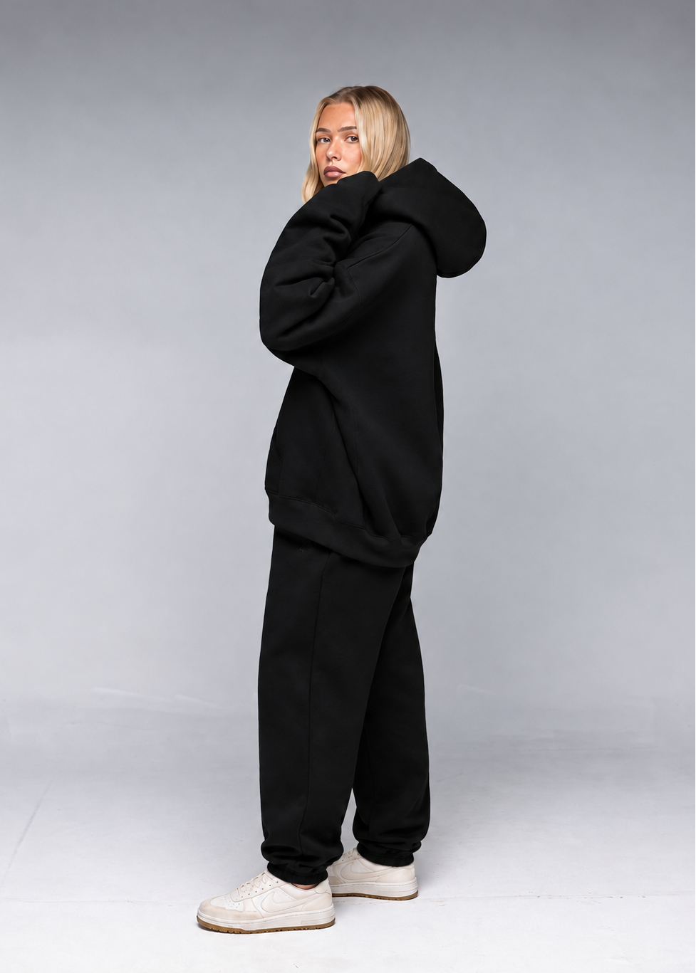 A woman modeling a black oversized hoodie and matching black sweatpants, paired with off-white sneakers, against a neutral gray studio background.
