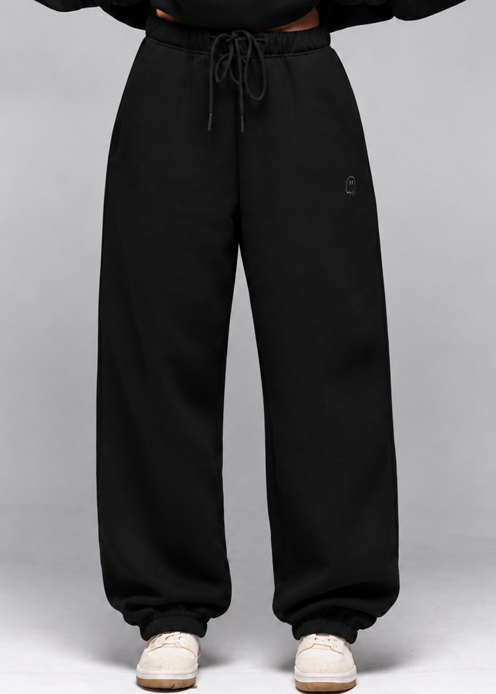 A model wearing black sweatpants with a drawstring waist and a matching black sweatshirt, paired with white sneakers, against a light gray studio background.