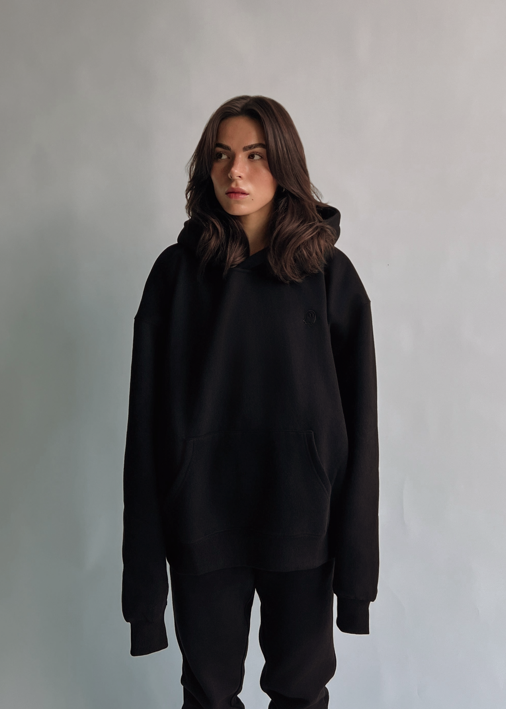 A woman with shoulder-length brown hair stands against a plain light gray wall wearing an oversized black hoodie and black pants.