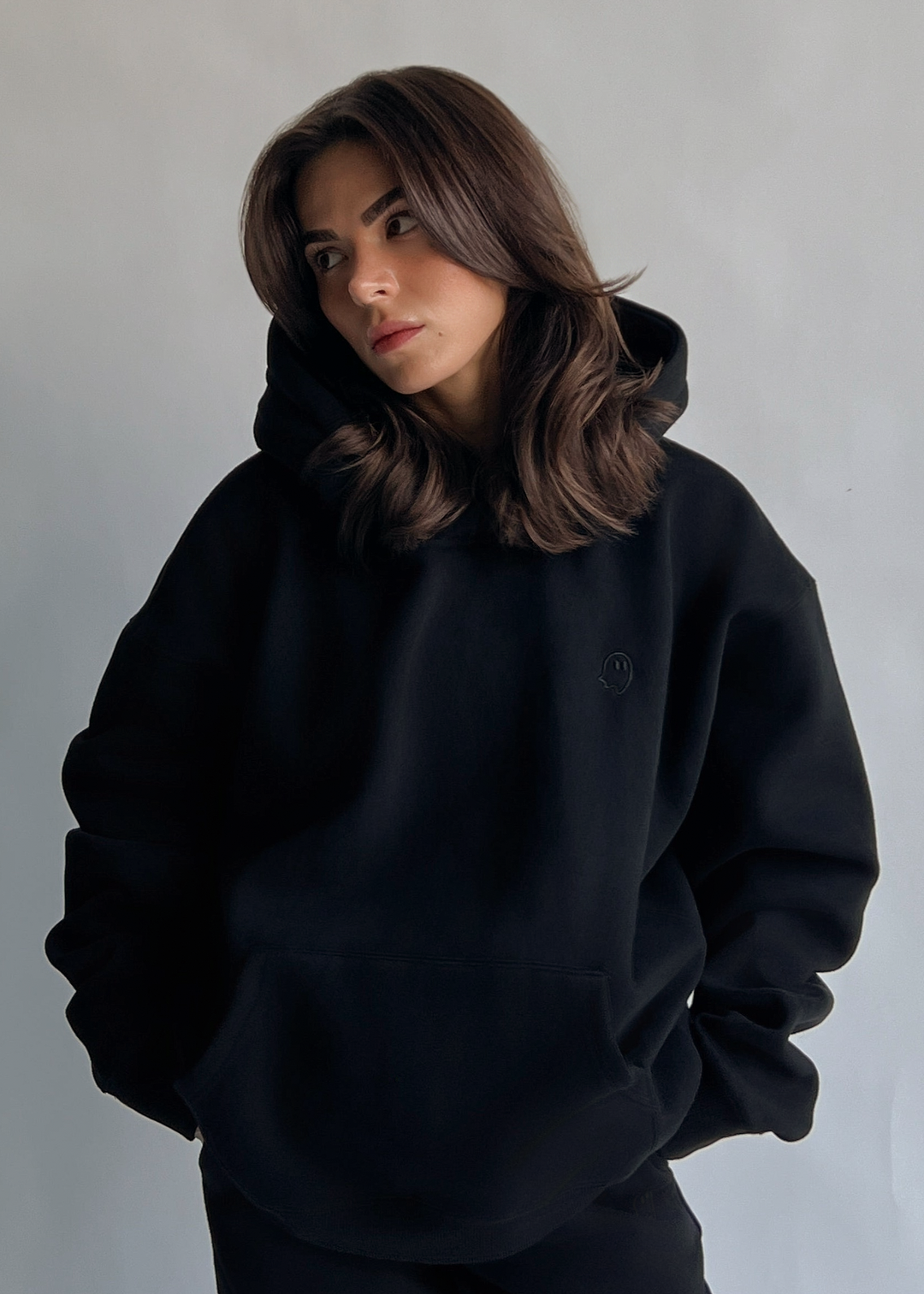 A woman with shoulder-length brown hair wearing a black oversized pullover hoodie and matching black pants, standing against a plain light gray wall.