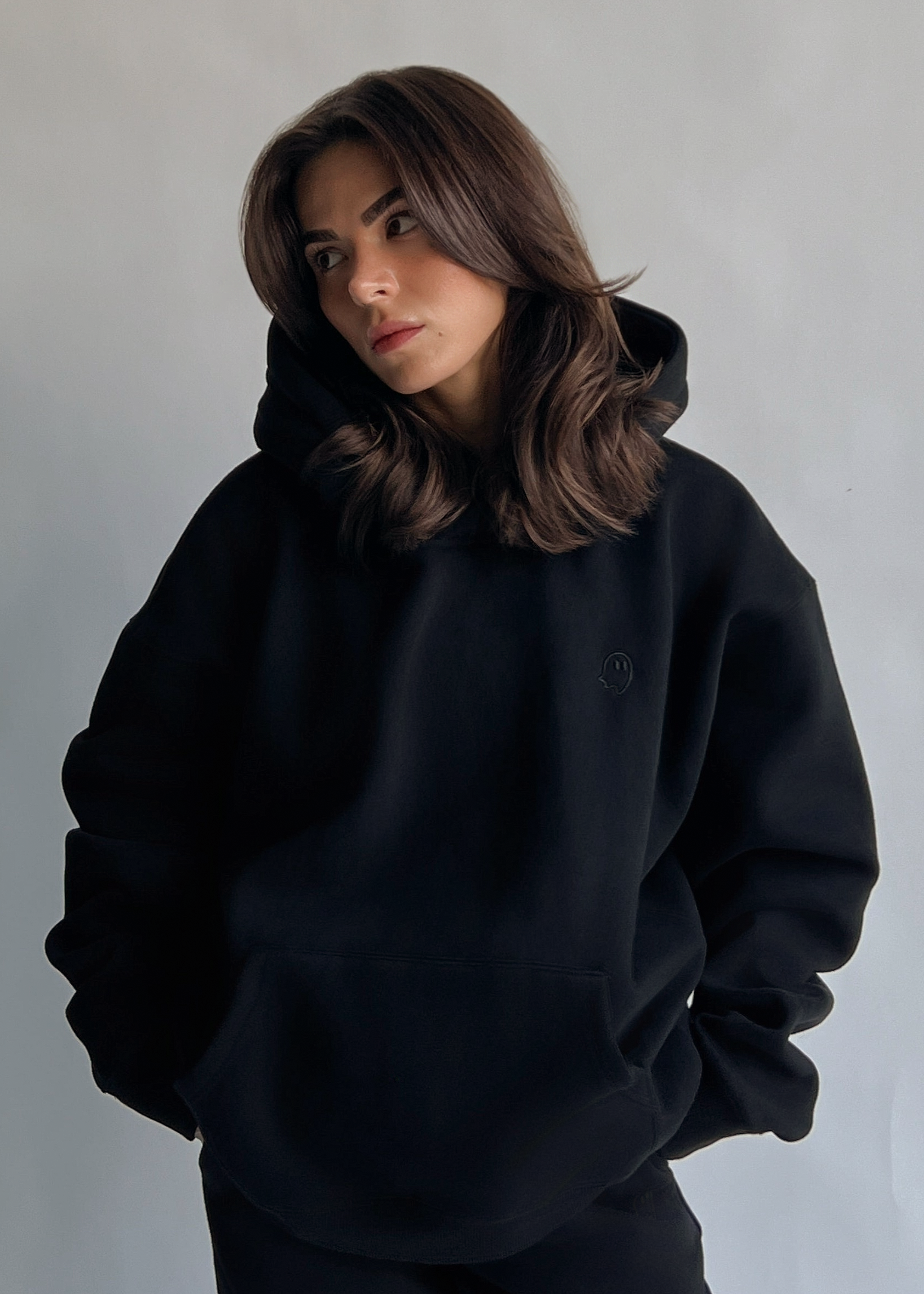 A woman with shoulder-length brown hair wearing a black oversized pullover hoodie and matching black pants, standing against a plain light gray wall.