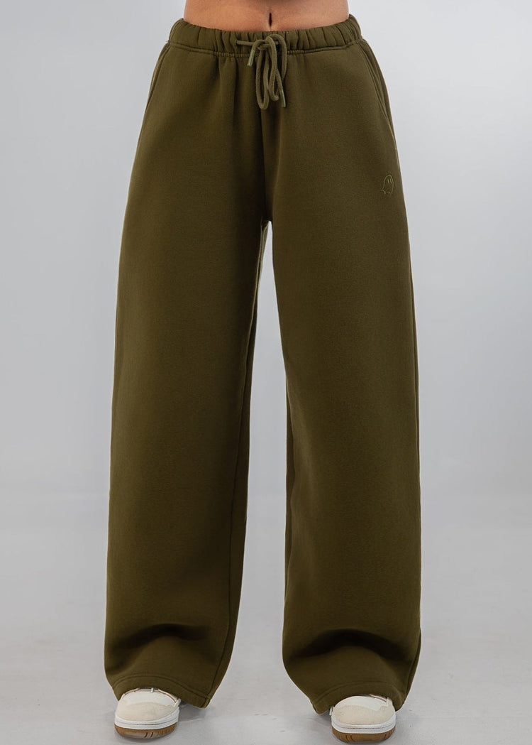 A model wearing olive green wide-leg sweatpants with a drawstring waist, paired with a matching cropped sweatshirt and white sneakers, shown from the waist down against a light gray studio background.