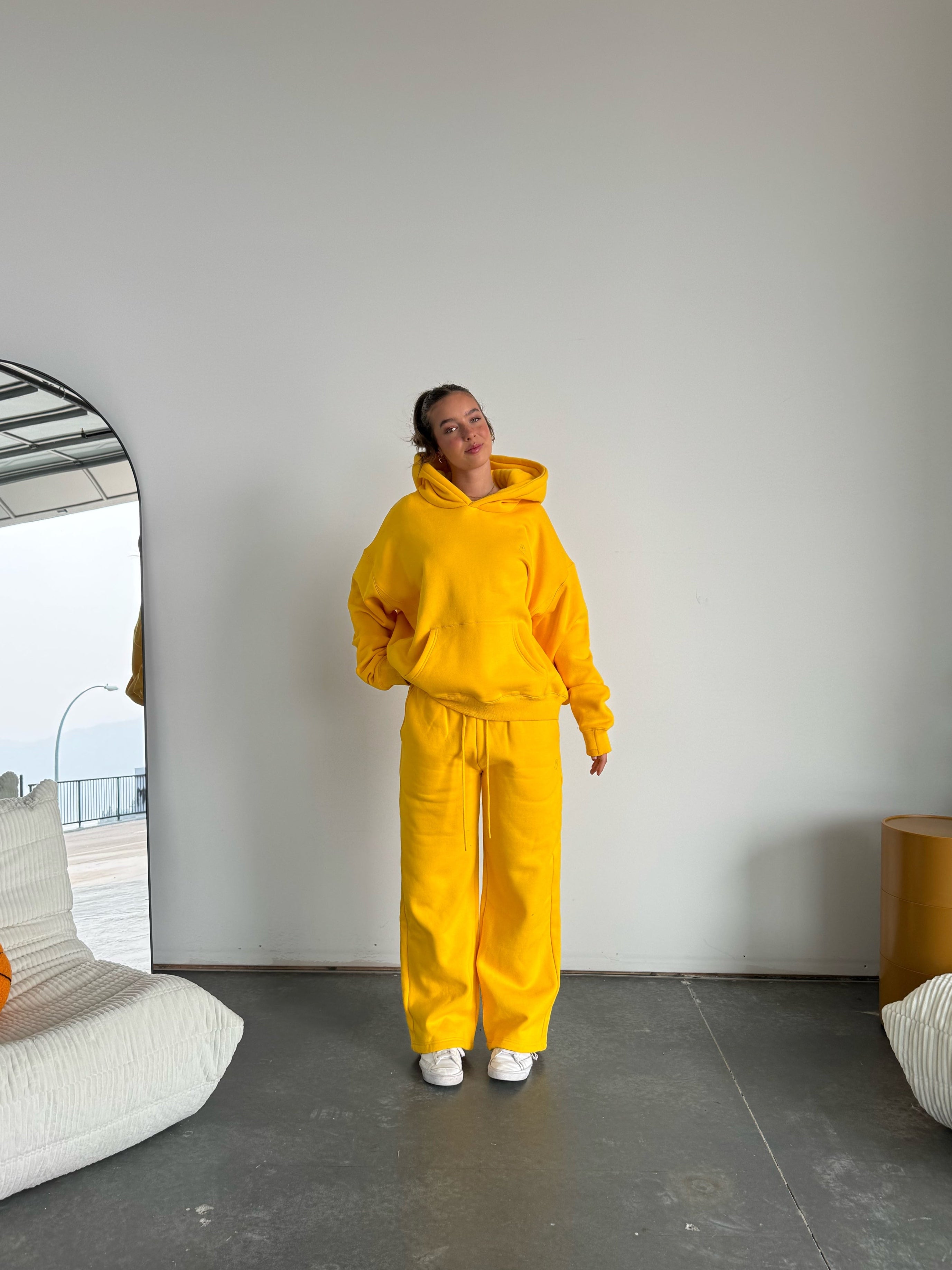 Person wearing a yellow hoodie and pants standing in a modern room with a white wall and gray floor.