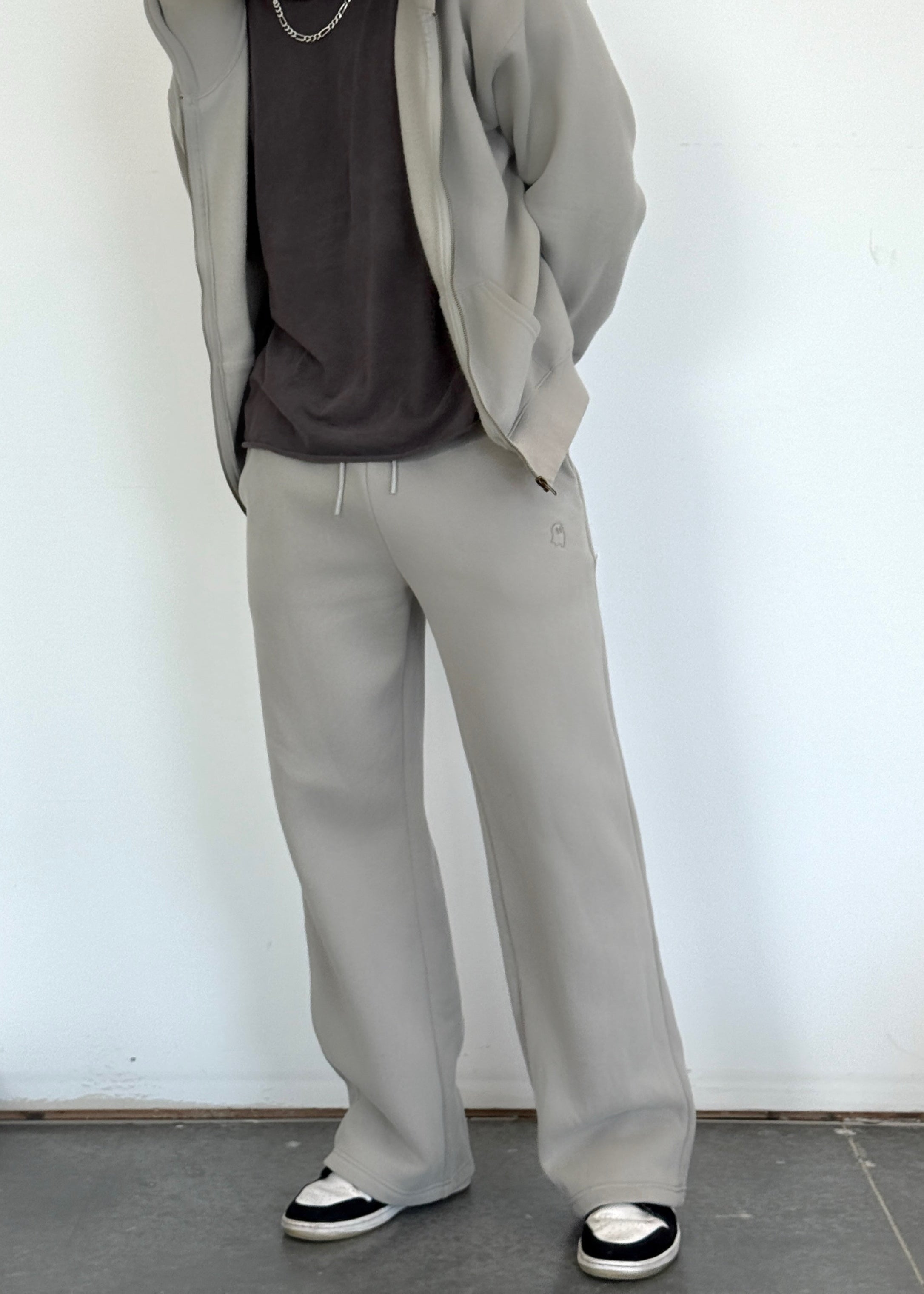 Person wearing a gray hoodie, dark shirt, light gray pants, and black cap against a white wall.