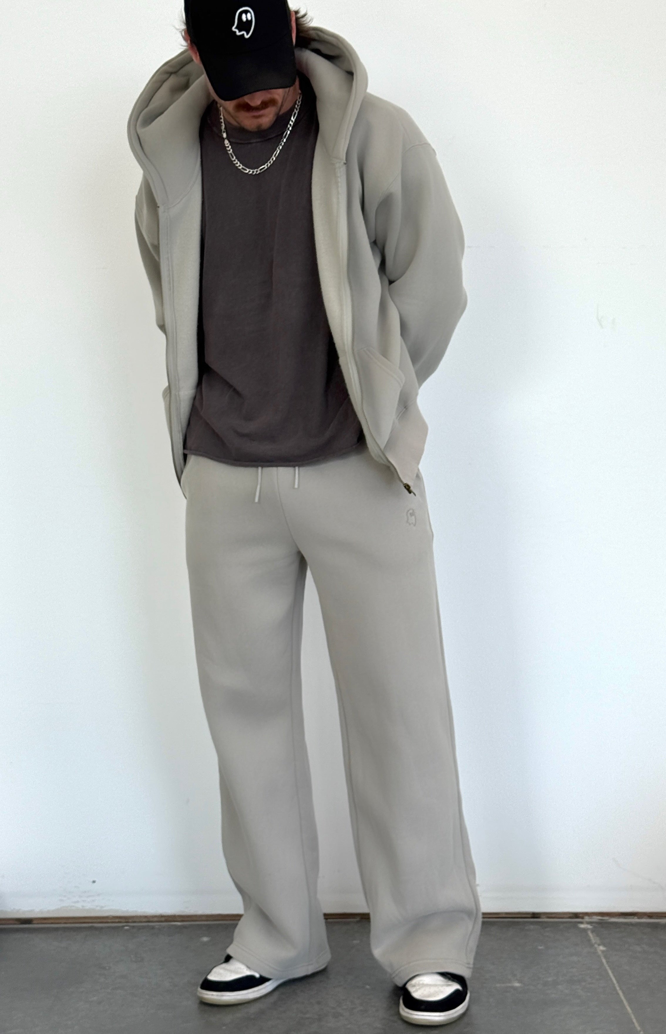 Person wearing a gray hoodie, gray pants, and black cap against a white wall.