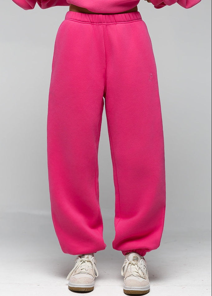 Bright pink sweatpants worn by a model, paired with a matching pink sweatshirt and white sneakers, shown from the waist down against a light gray studio background.