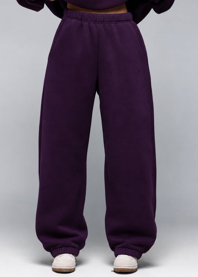 A model wearing a matching deep purple sweatshirt and wide-leg sweatpants set, shown from the neck down against a neutral gray studio background.
