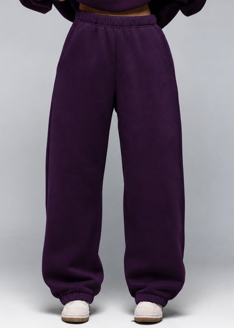 A model wearing a matching deep purple sweatshirt and wide-leg sweatpants set, shown from the neck down against a neutral gray studio background.