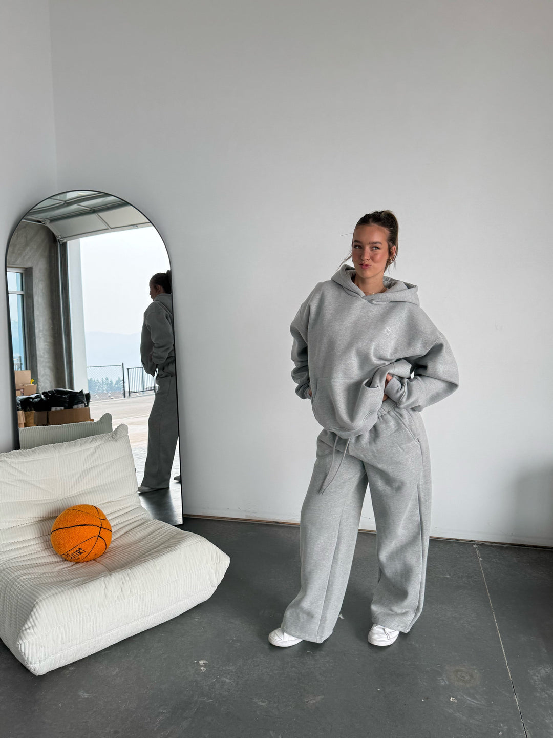 Person in gray hoodie and pants standing in a minimalistic room with a basketball and white cushion.