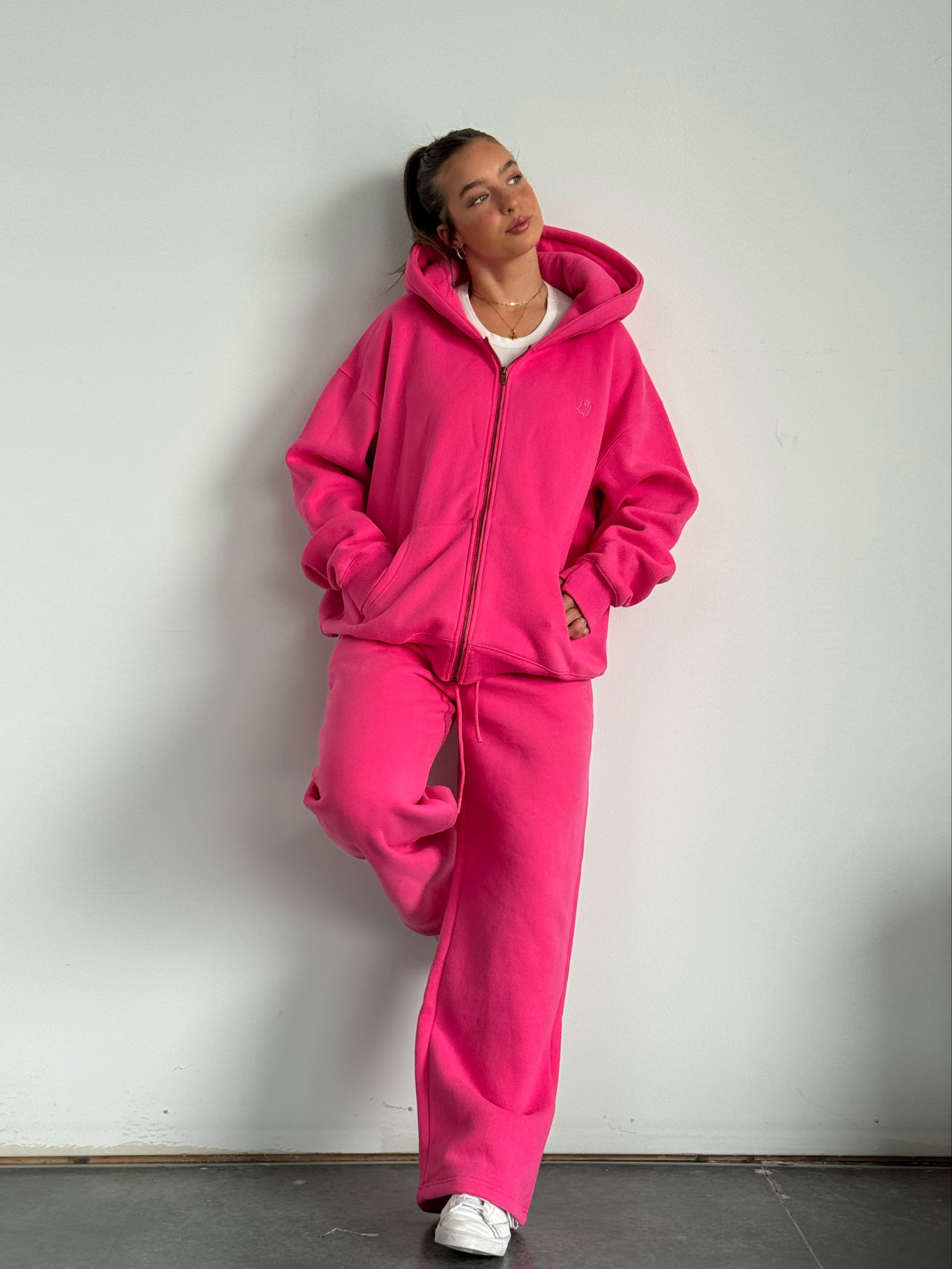 Person wearing a bright pink hoodie and pants against a white wall.