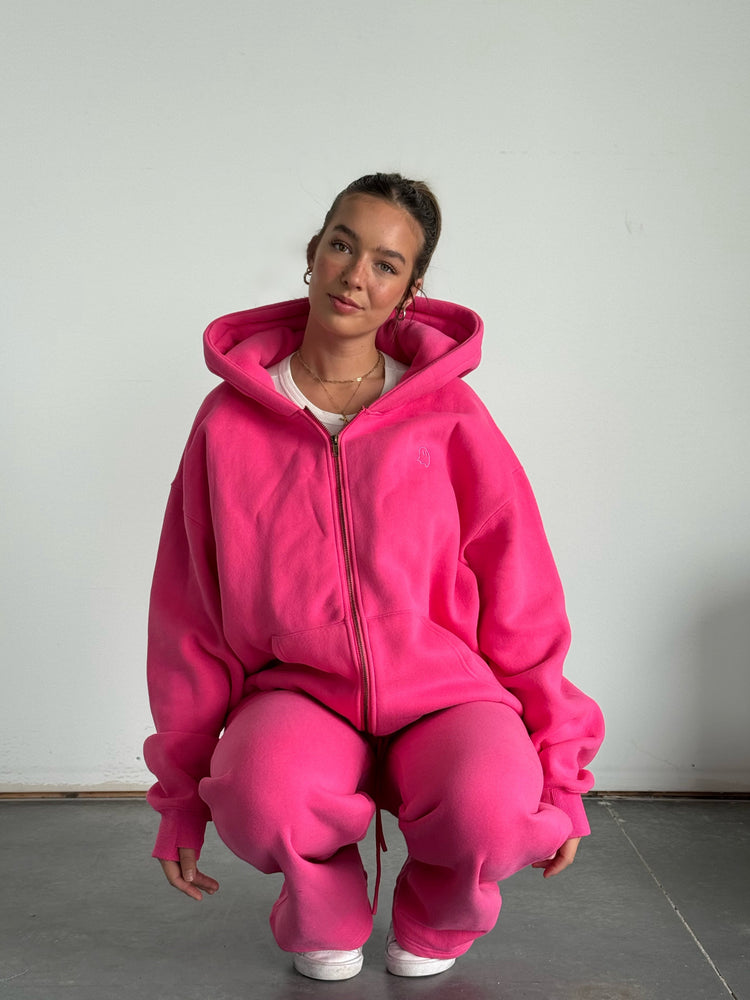 Person wearing a bright pink hoodie and pants sitting on the floor against a plain background
