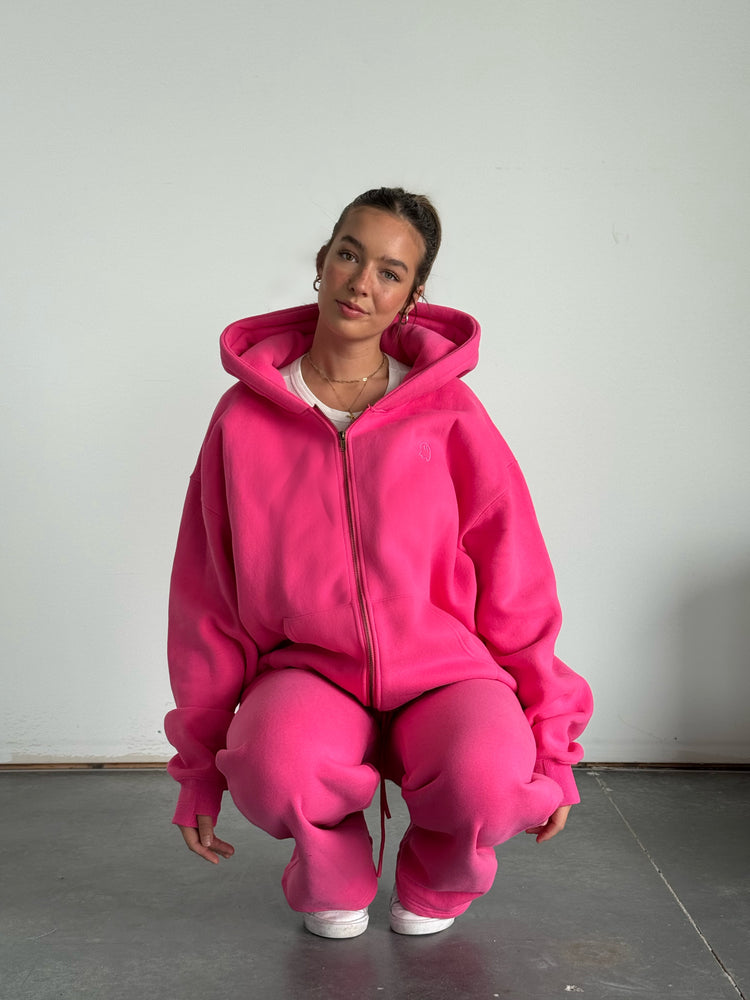 Person wearing a bright pink hoodie and pants sitting on the floor against a plain background