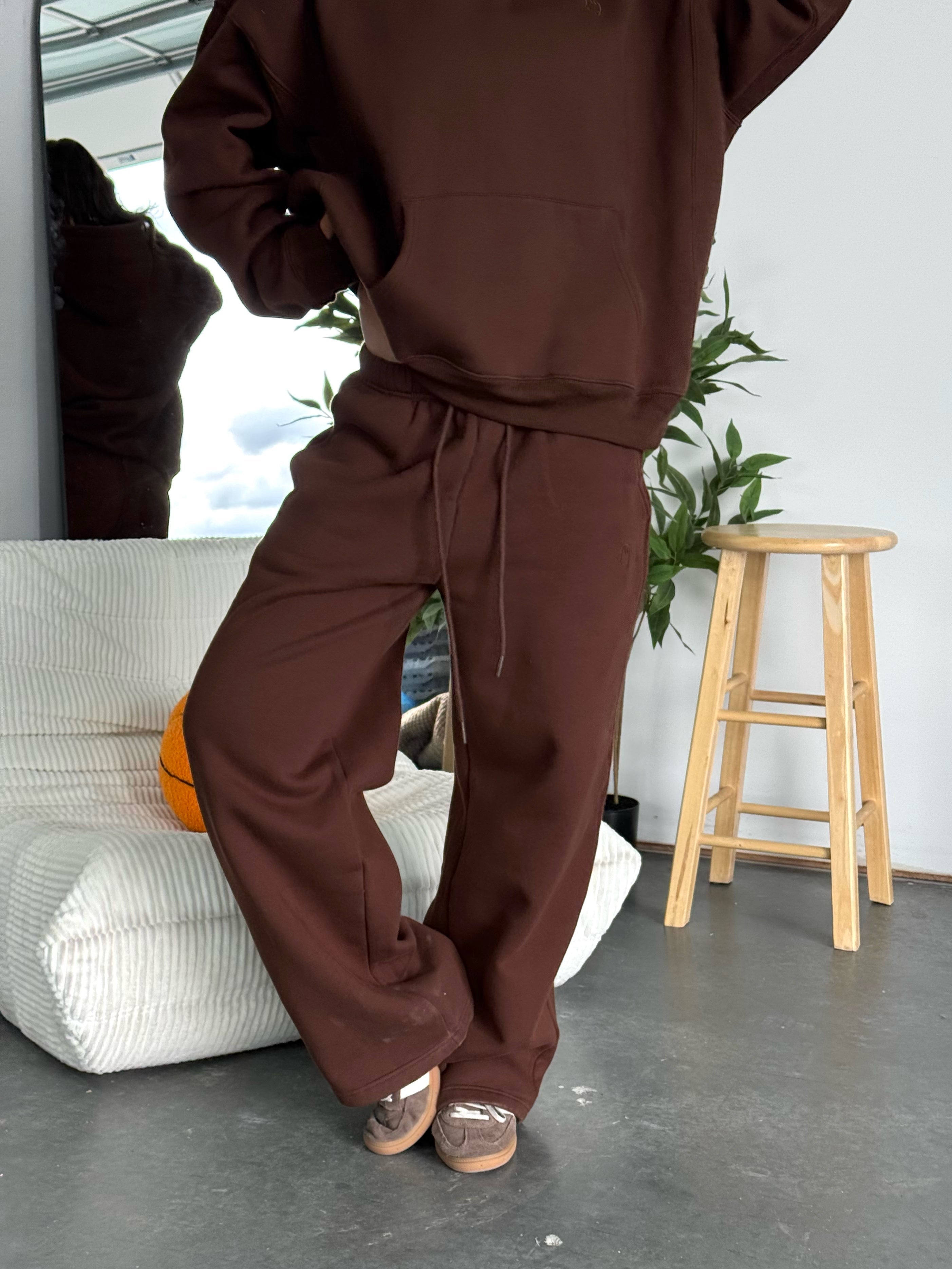 Person wearing a brown tracksuit in a room with a stool and plant.