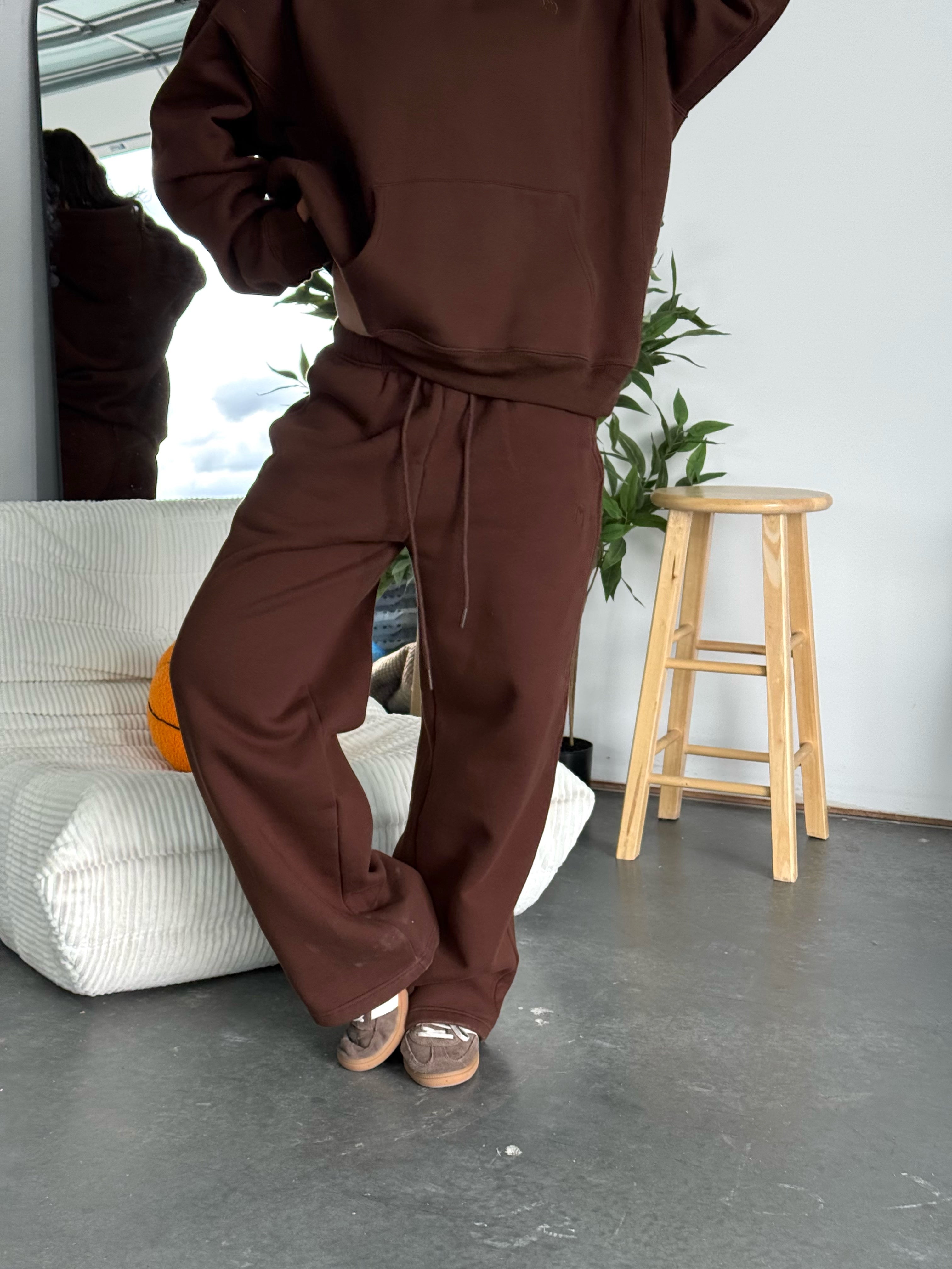 Person wearing a brown tracksuit in a room with a stool and plant.