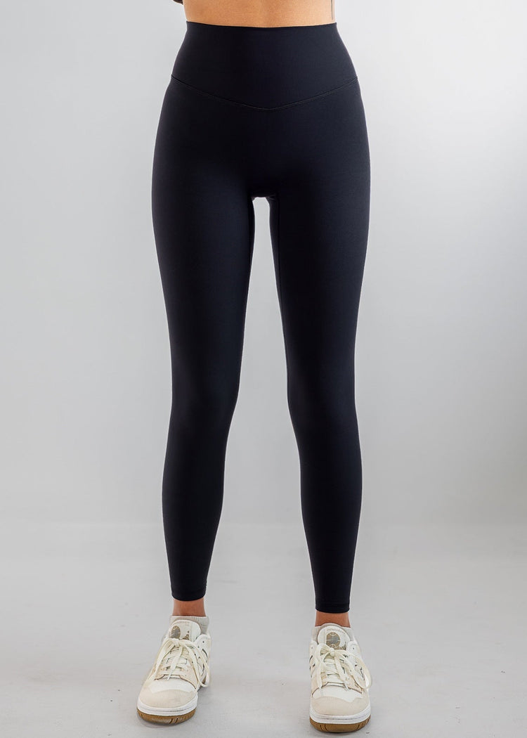 A model from the waist down wearing high-waisted black leggings, a brown cropped sweatshirt, and white sneakers, standing against a light gray studio background.