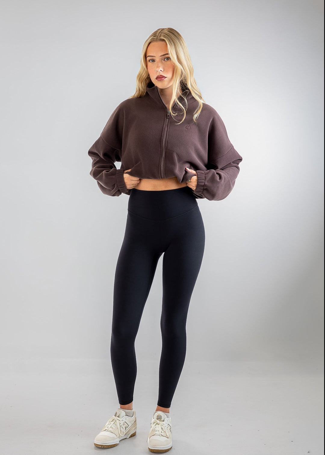 A woman modeling a dark brown cropped zip-up sweatshirt and black leggings with white sneakers, standing against a light gray studio background.
