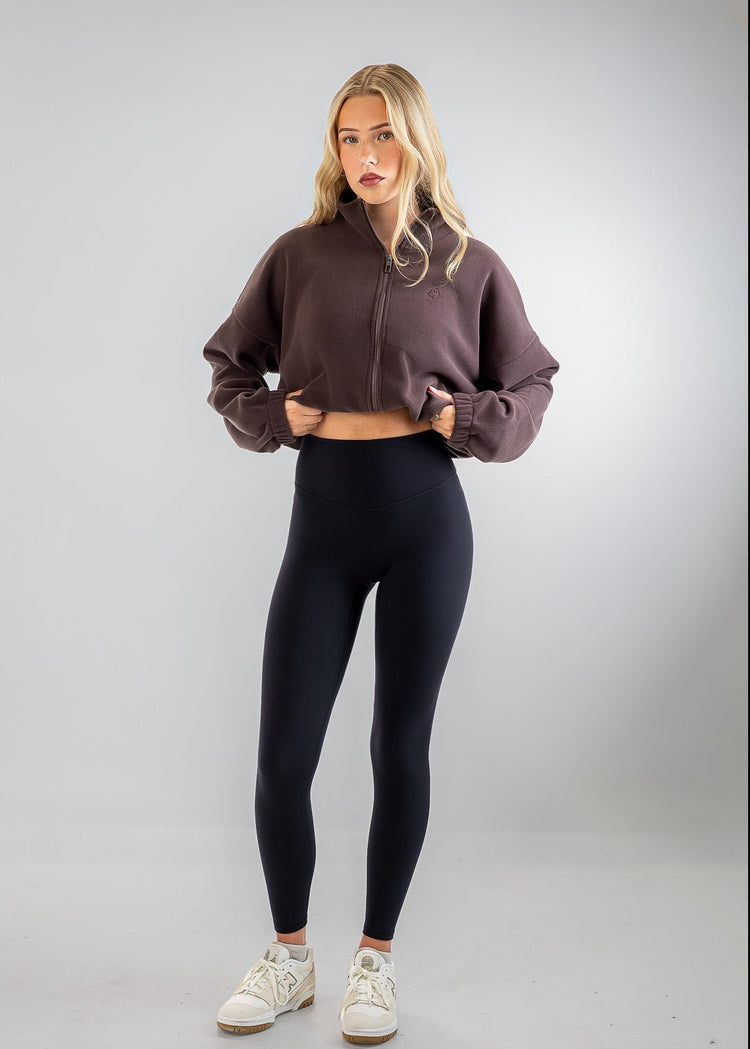 A woman modeling a dark brown cropped zip-up sweatshirt and black leggings with white sneakers, standing against a light gray studio background.