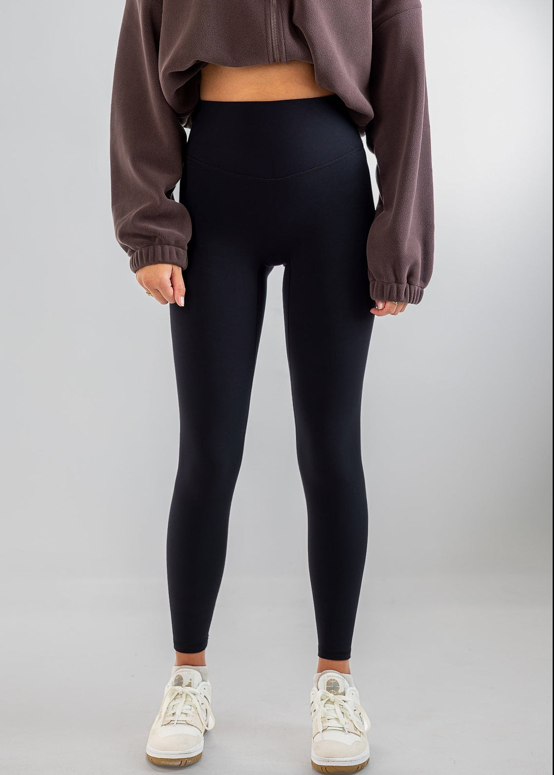A model wearing high-waisted black leggings, a brown cropped sweatshirt, and white sneakers, standing against a light gray studio background.