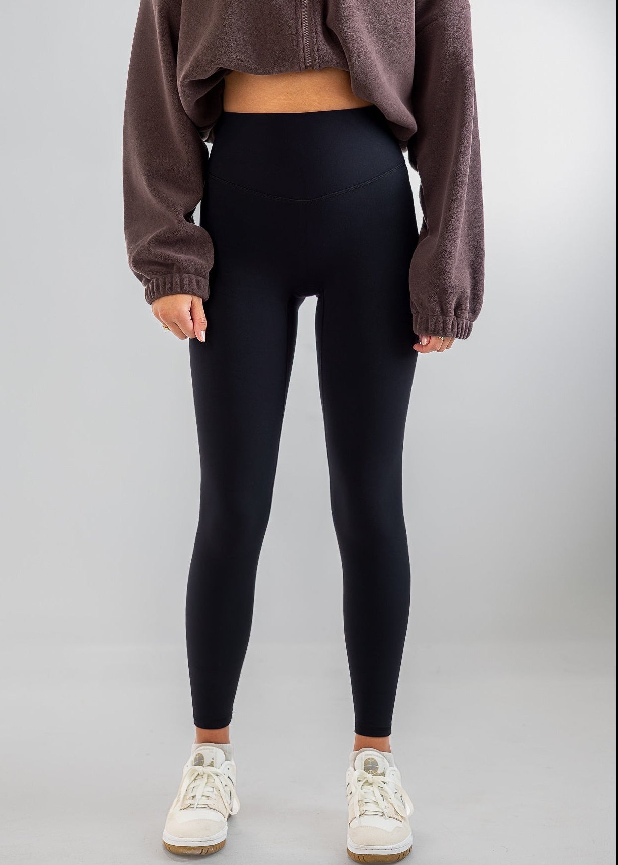 A model wearing high-waisted black leggings, a brown cropped sweatshirt, and white sneakers, standing against a light gray studio background.