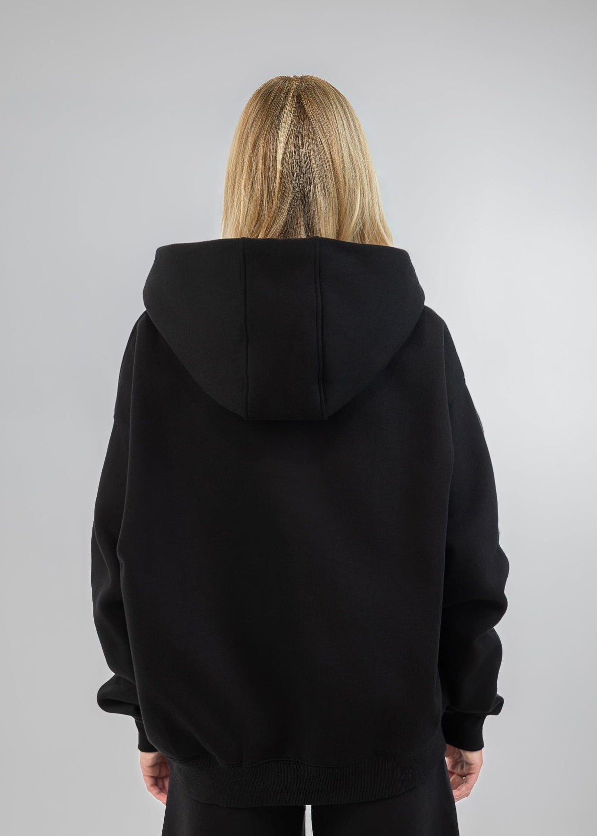A woman viewed from the back wearing an oversized black hoodie and black pants against a light gray studio background.