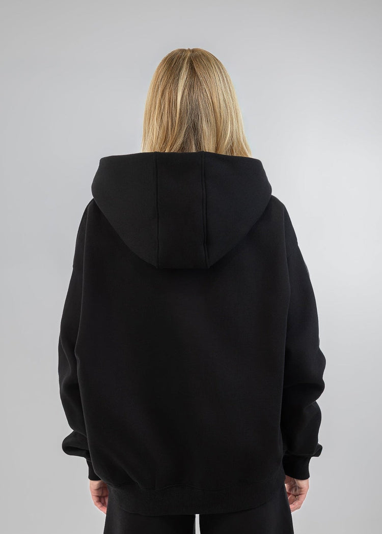 A woman viewed from the back wearing an oversized black hoodie and black pants against a light gray studio background.