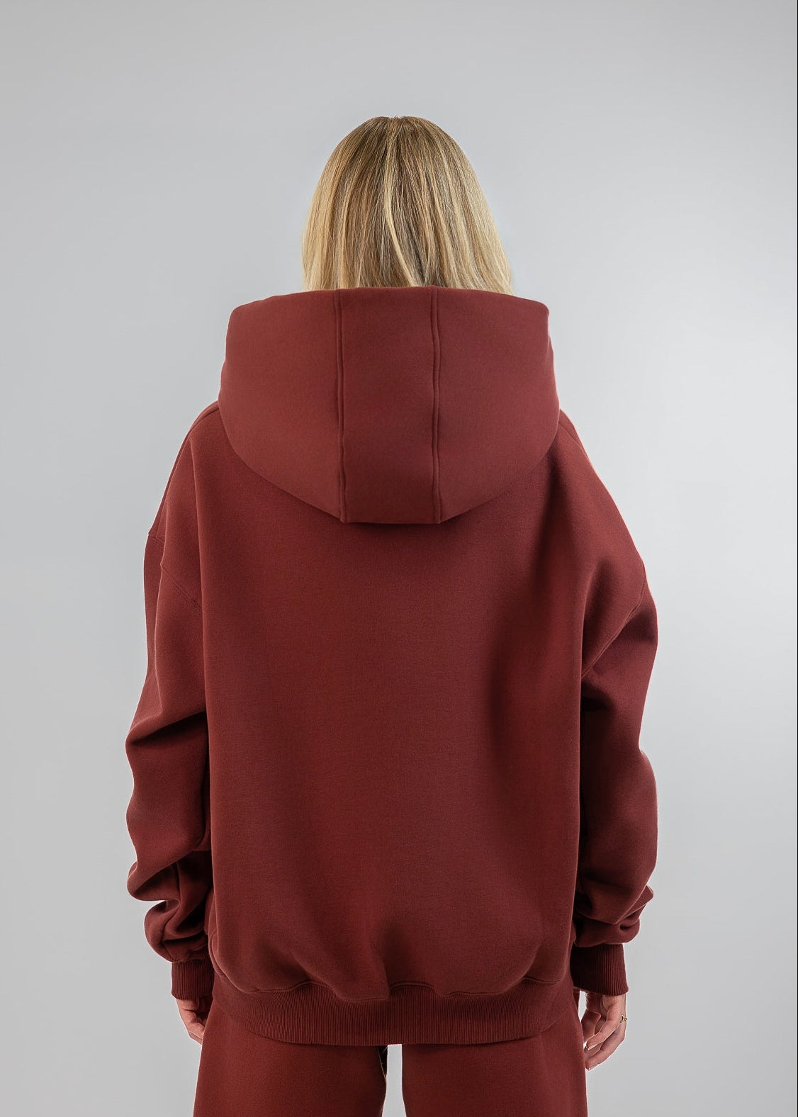 A model viewed from behind wearing a matching rust-red hoodie and sweatpants set against a plain light gray studio background.