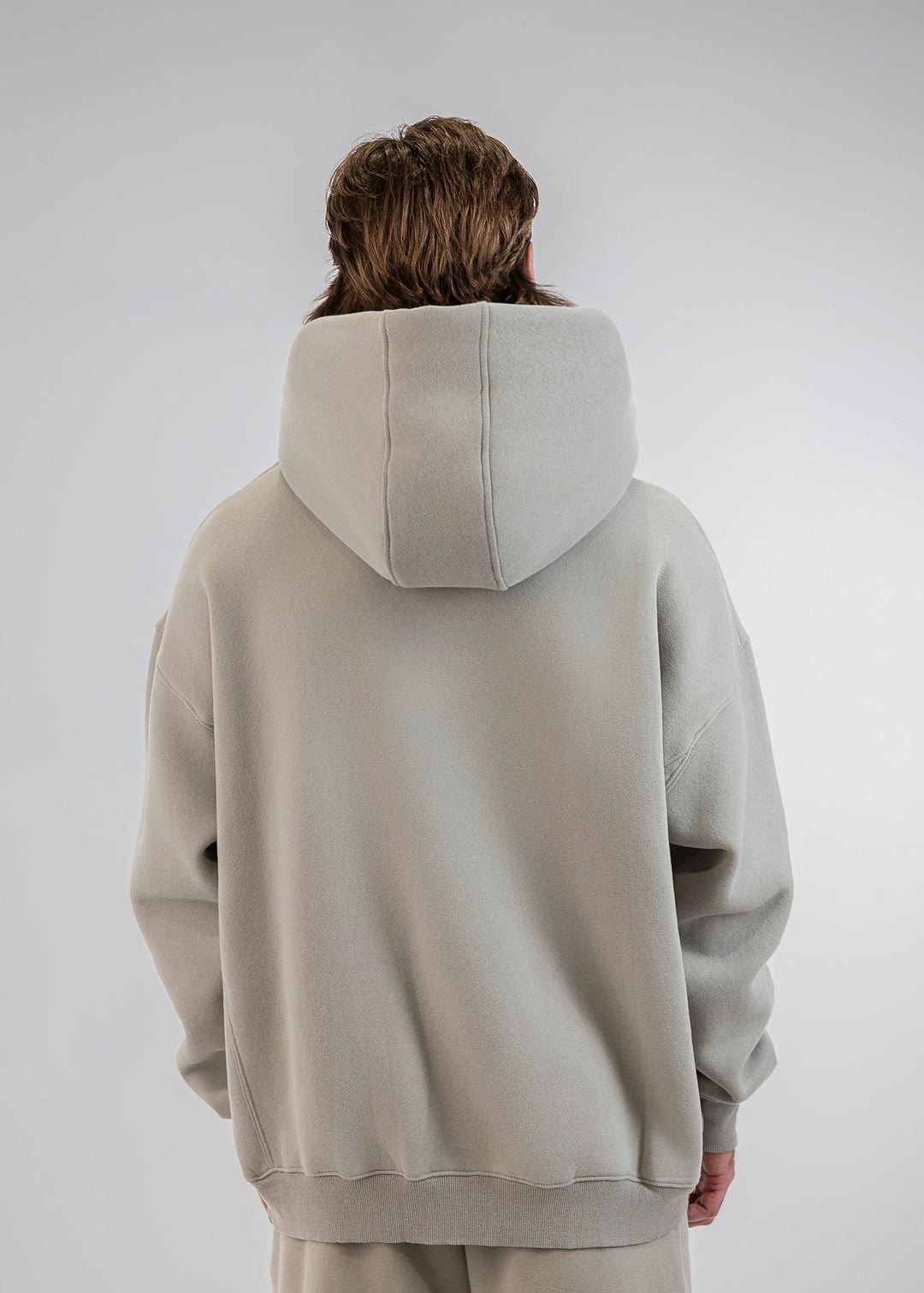 A person viewed from behind wearing an oversized light beige hoodie and matching sweatpants against a plain light gray studio background.