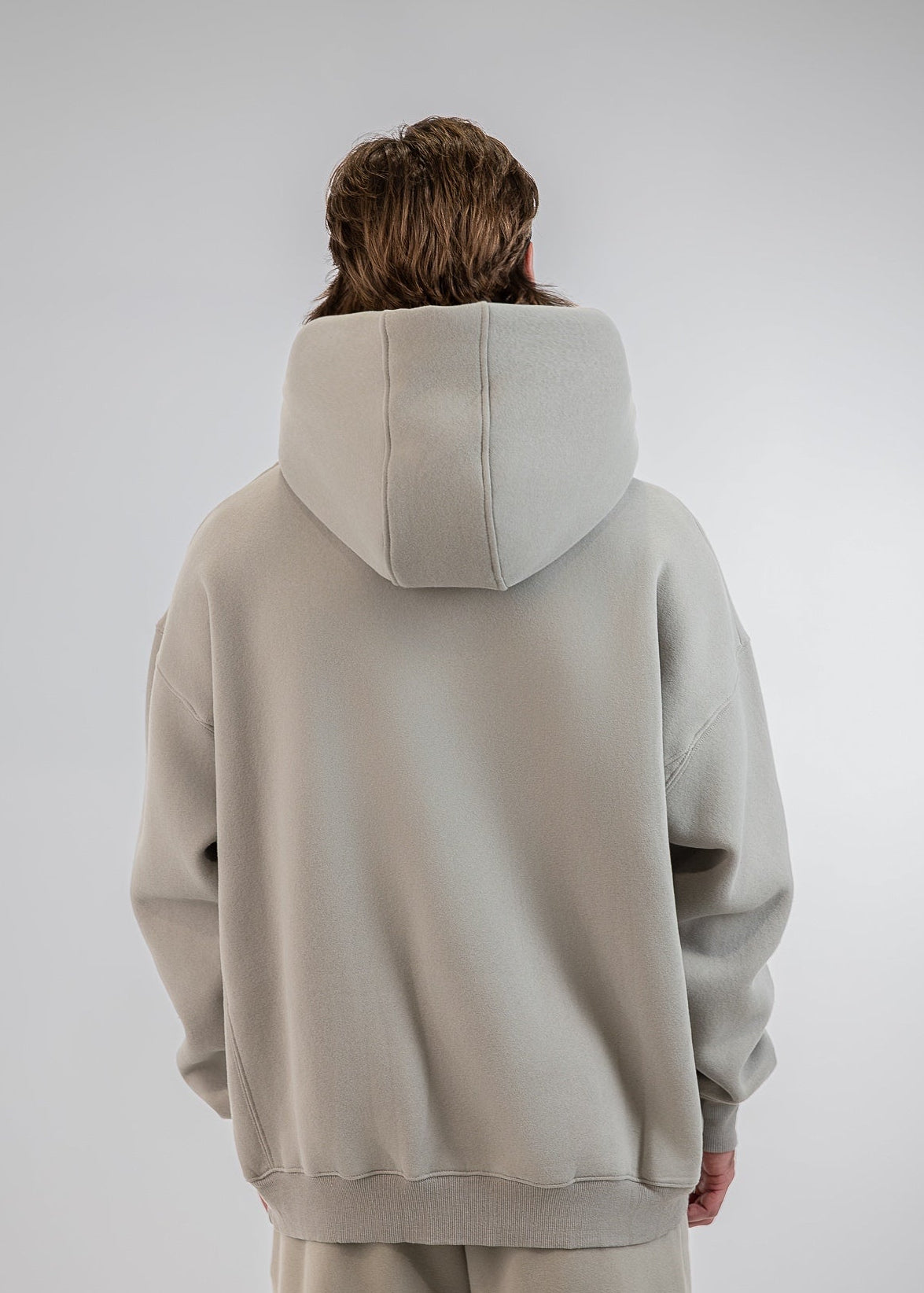 A person viewed from behind wearing an oversized light beige hoodie and matching sweatpants against a plain light gray studio background.