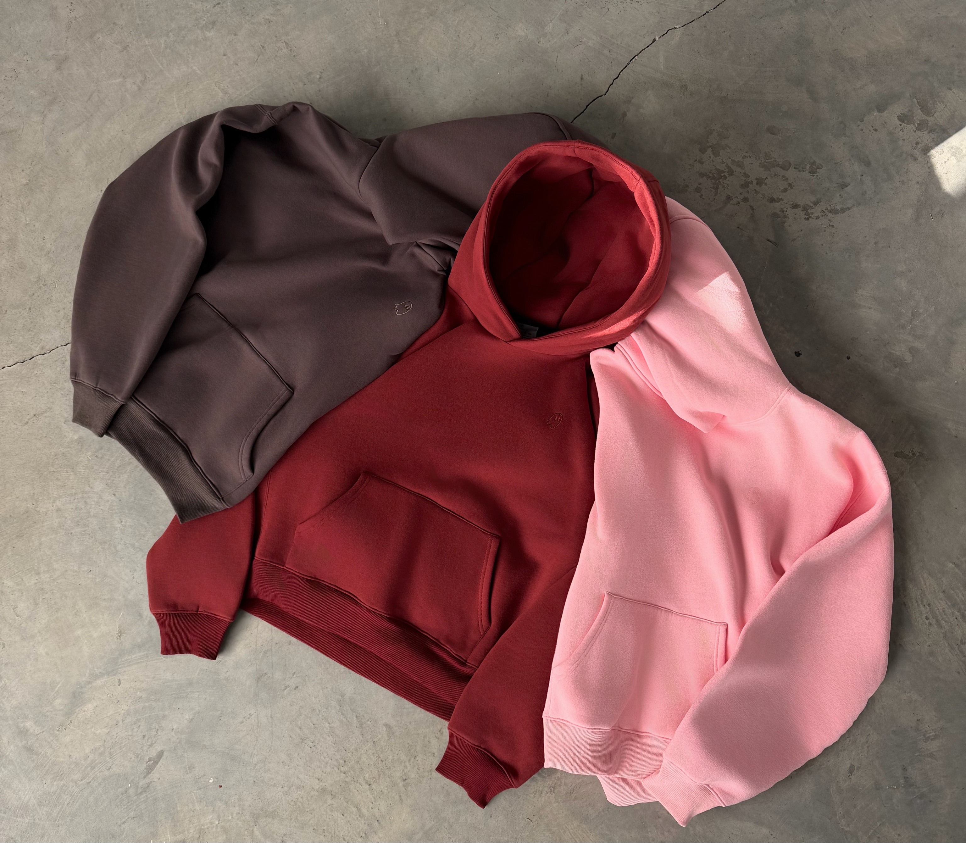 Three hoodies in brown, red, and pink on a gray concrete floor.