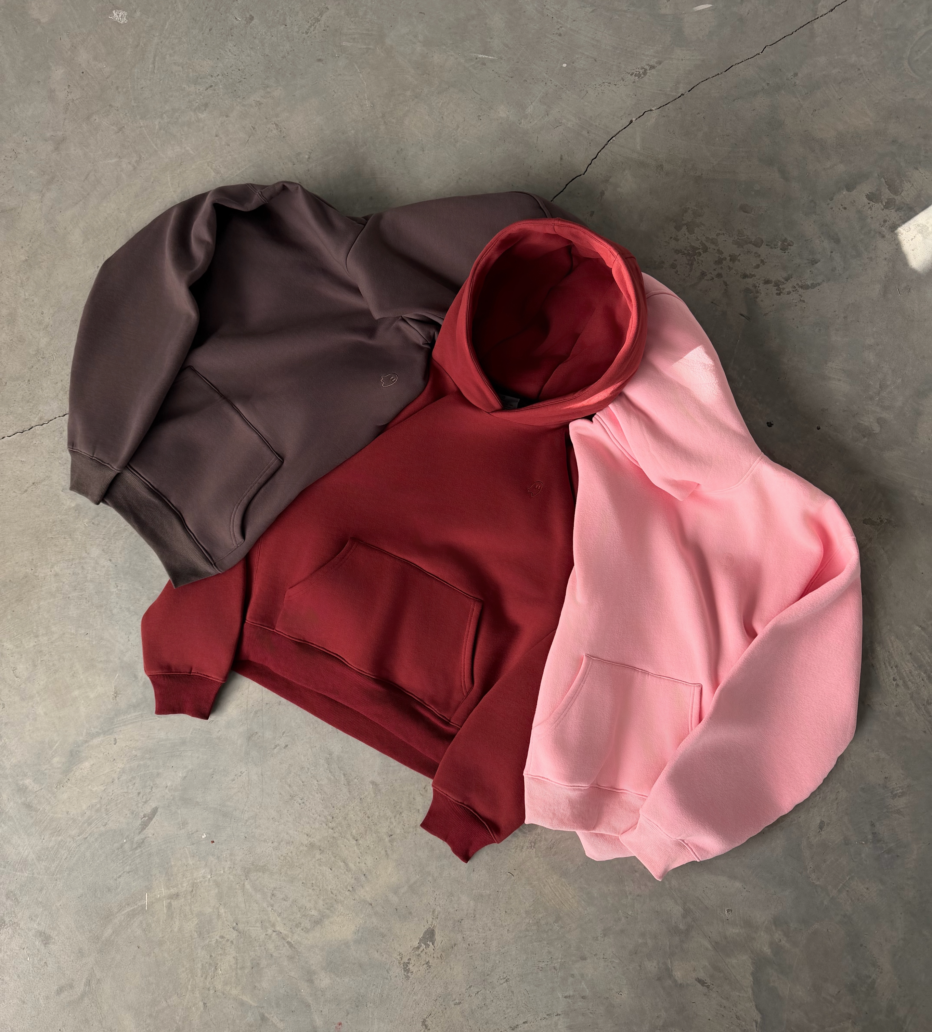 Three hoodies in brown, red, and pink on a gray concrete floor.