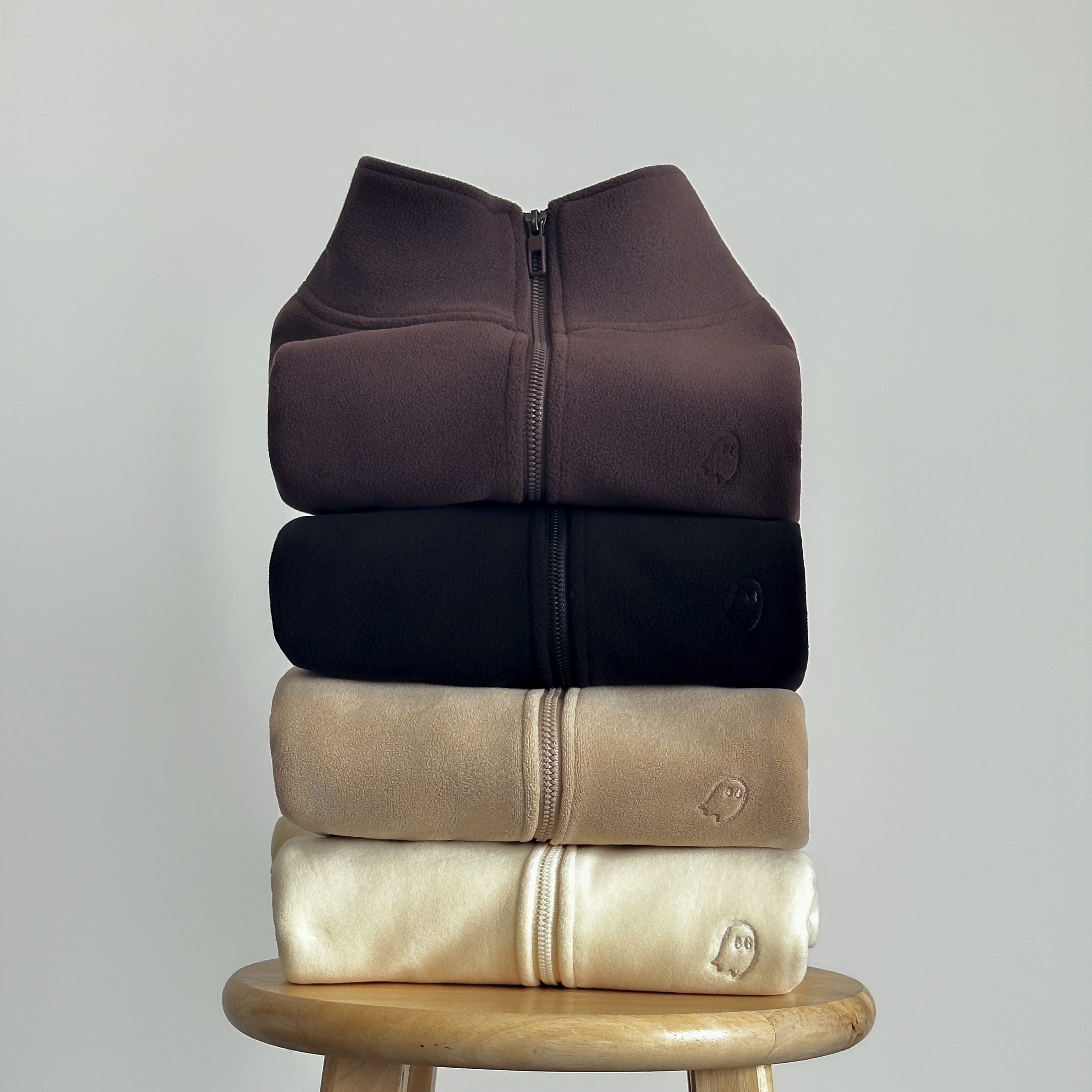 Four folded zip-front sweatshirts stacked vertically on a wooden stool, shown in dark brown, black, beige, and cream against a light gray background.
