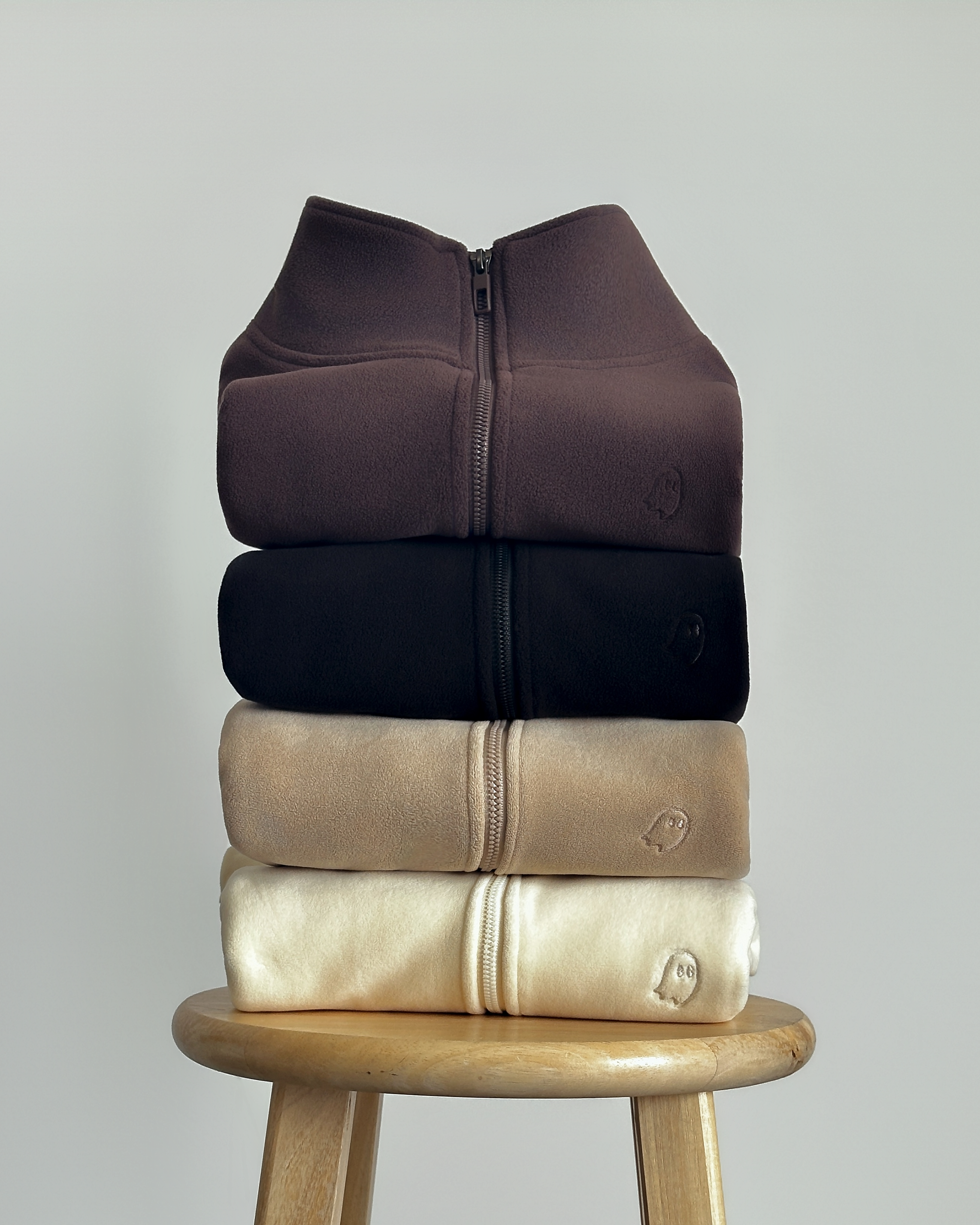 Four folded zip-front sweatshirts stacked vertically on a wooden stool, shown in dark brown, black, beige, and cream against a light gray background.