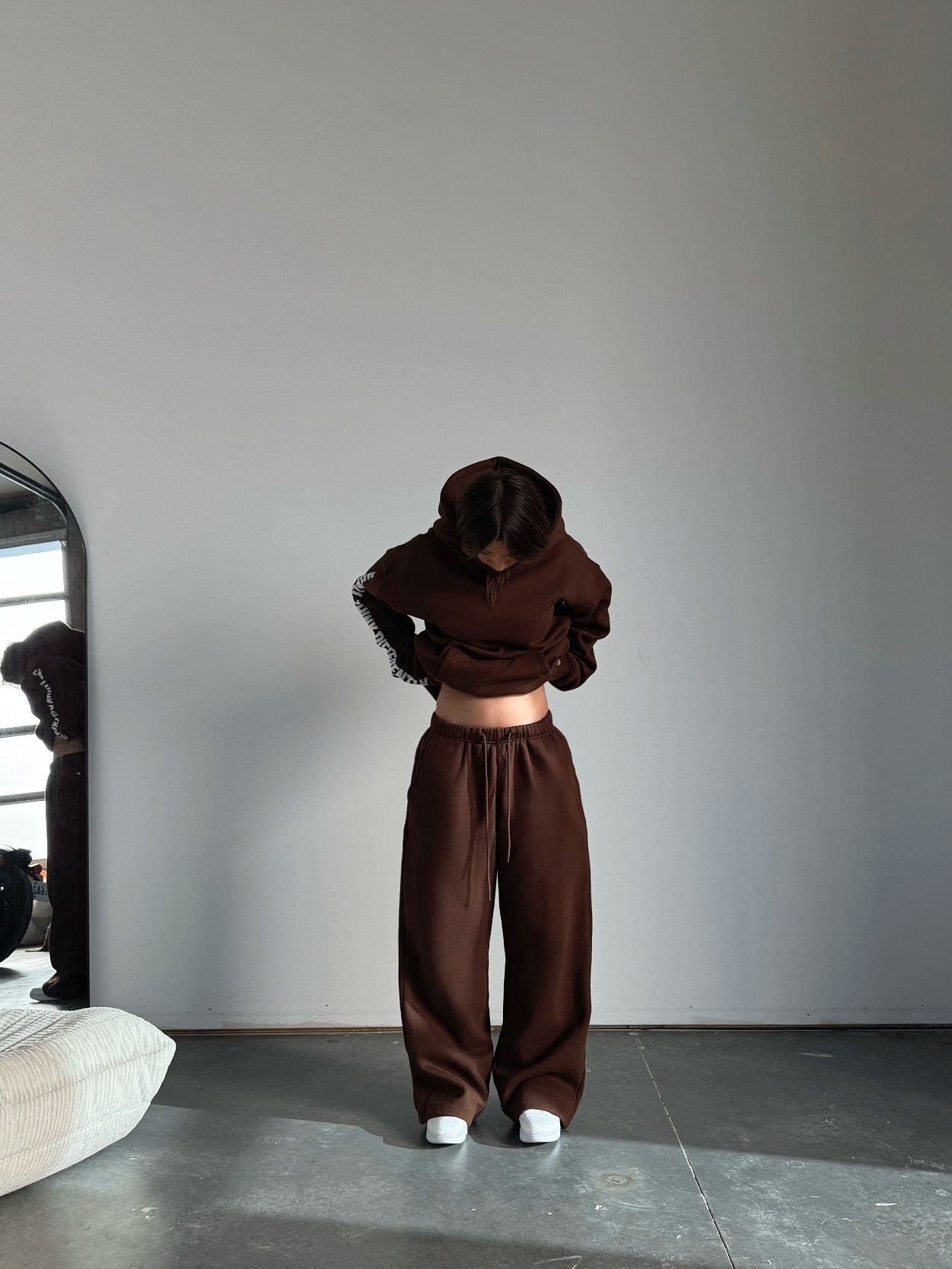 Person wearing a brown outfit standing in a minimalistic room with a mirror.