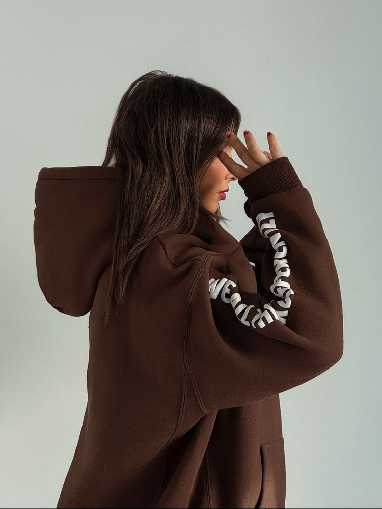 Person wearing a brown hoodie with white text on sleeves against a plain background
