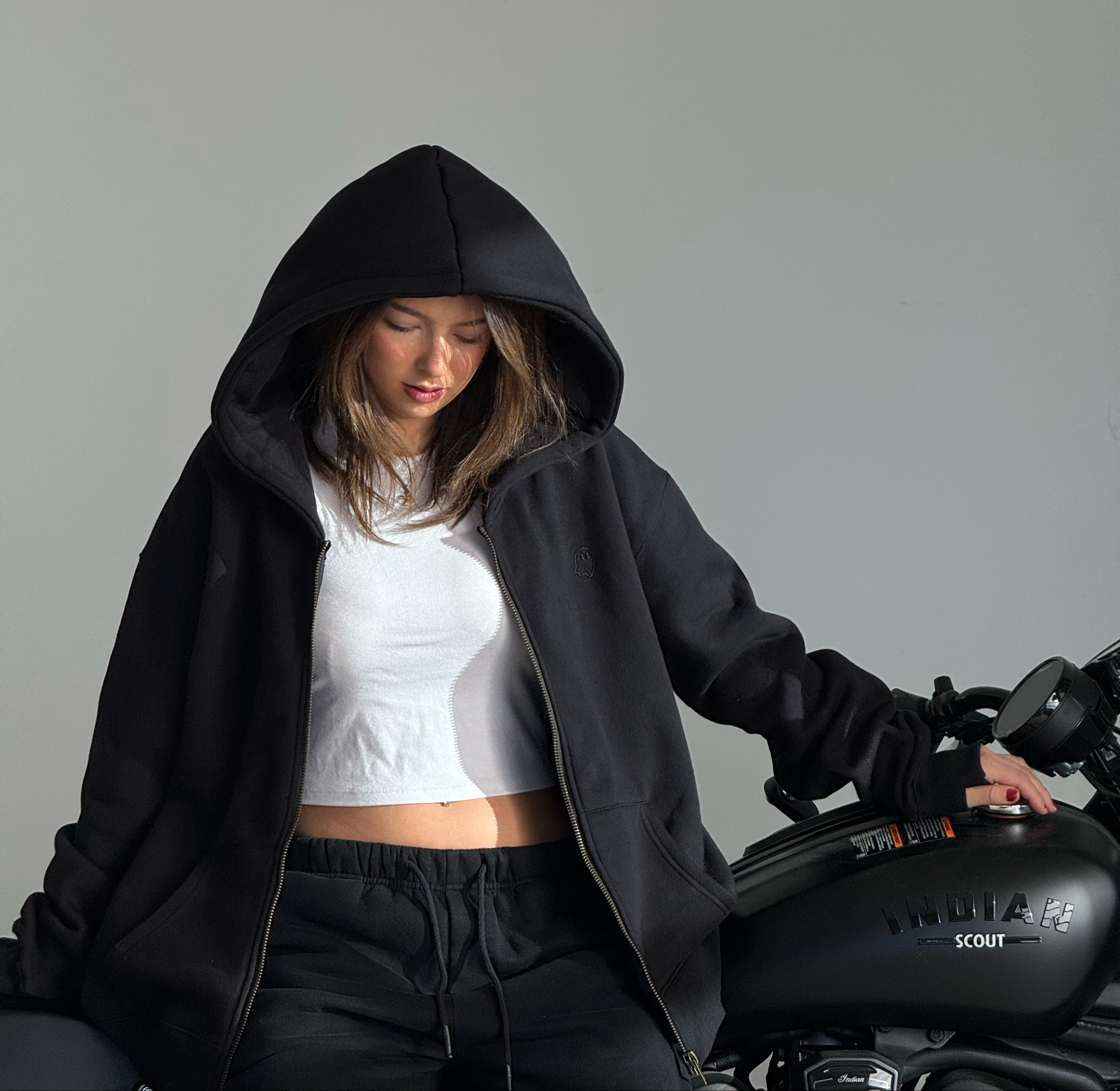 Person wearing a black hoodie and white top sitting next to a motorcycle on a gray background