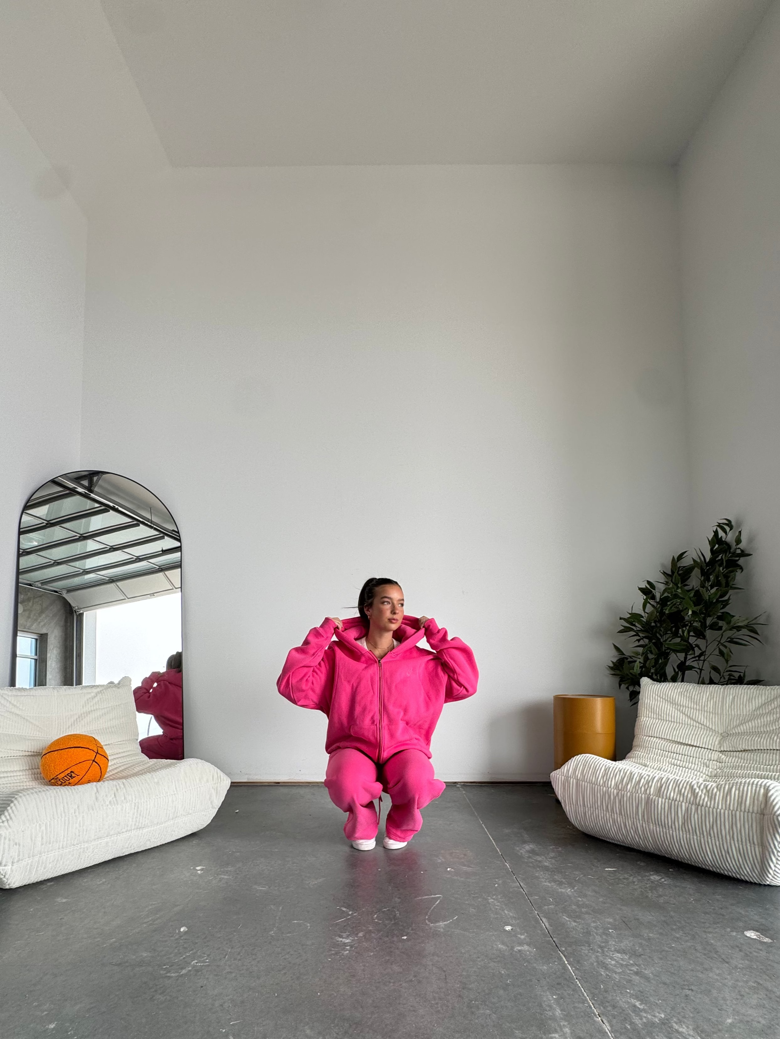 Person in a bright pink outfit sitting on a white chair in a minimalistic room with a mirror and plant.