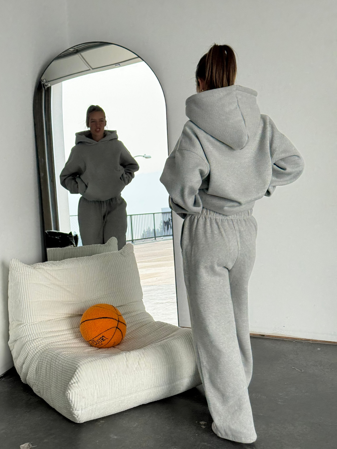 Person in a gray tracksuit standing in front of a mirror with a basketball on a white chair.