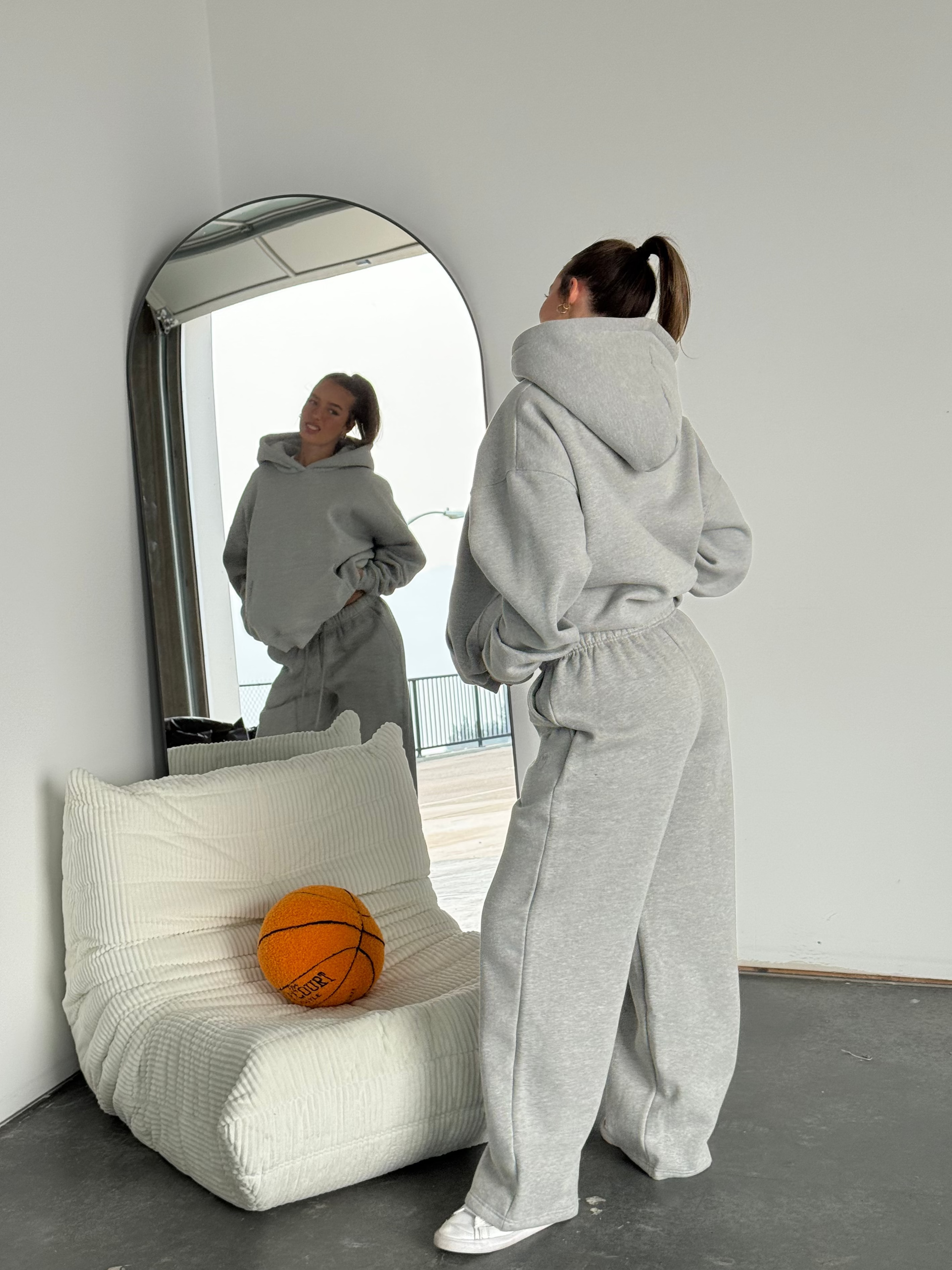 Person in a gray tracksuit standing in front of a mirror with a basketball on a white chair.