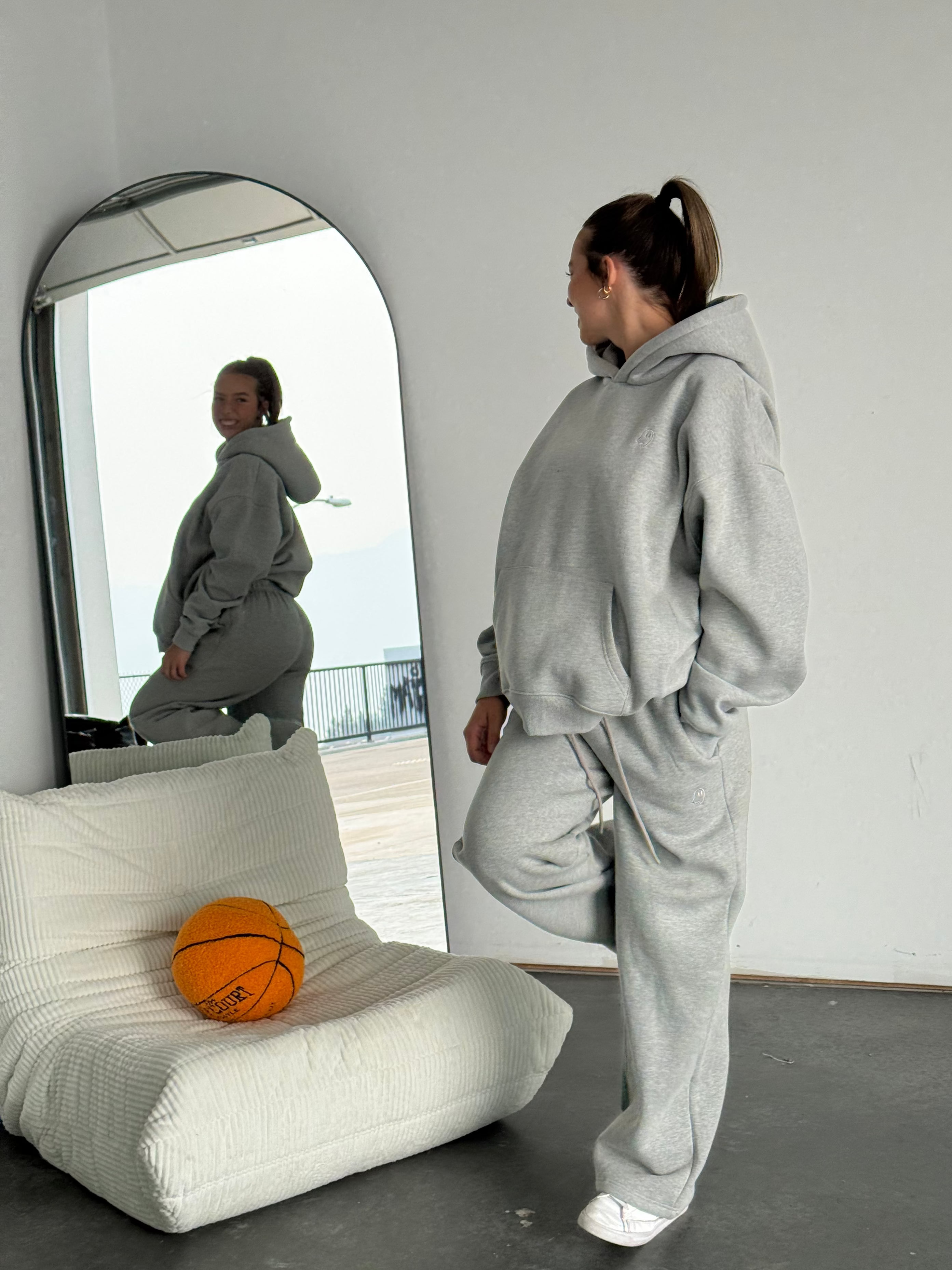 Person in a gray tracksuit standing in front of a mirror with a basketball on a white chair.