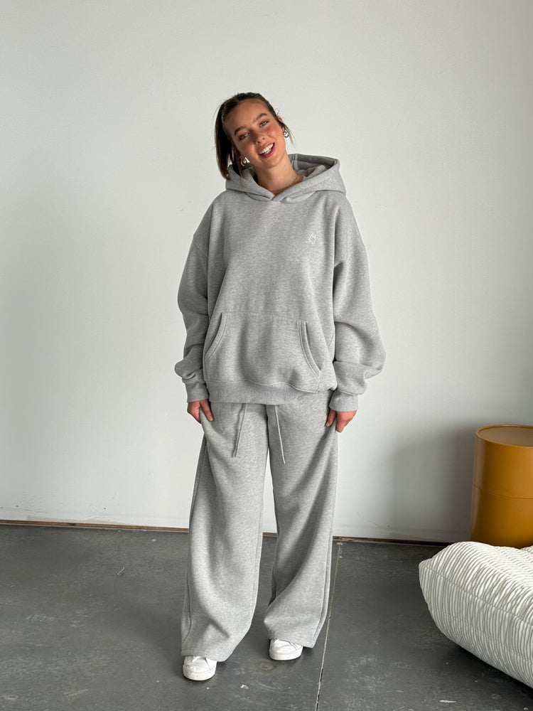 Person wearing a gray hoodie and pants standing in a minimalistic room with a white wall and gray floor.