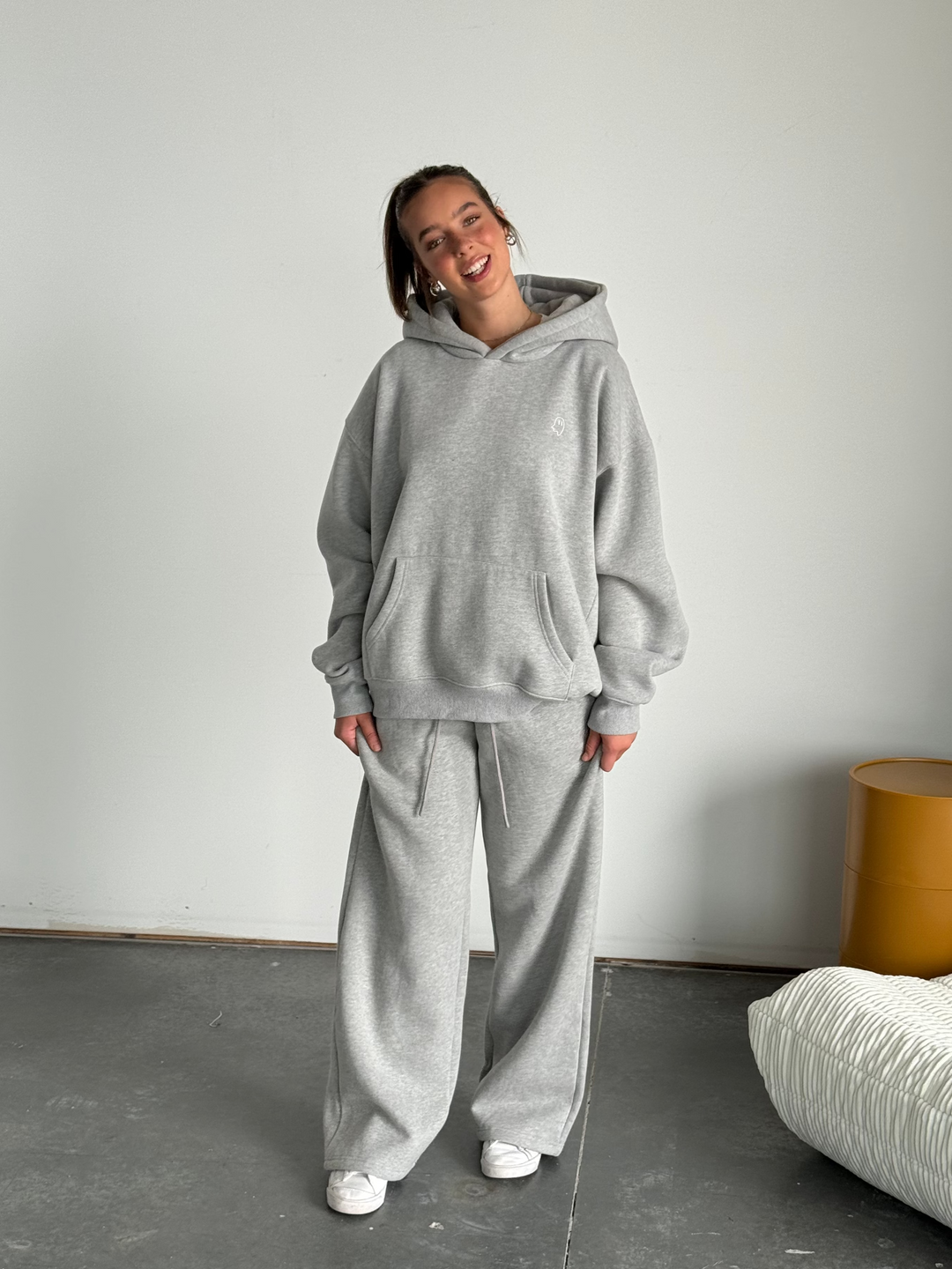 Person wearing a gray hoodie and pants standing in a minimalistic room with a white wall and gray floor.