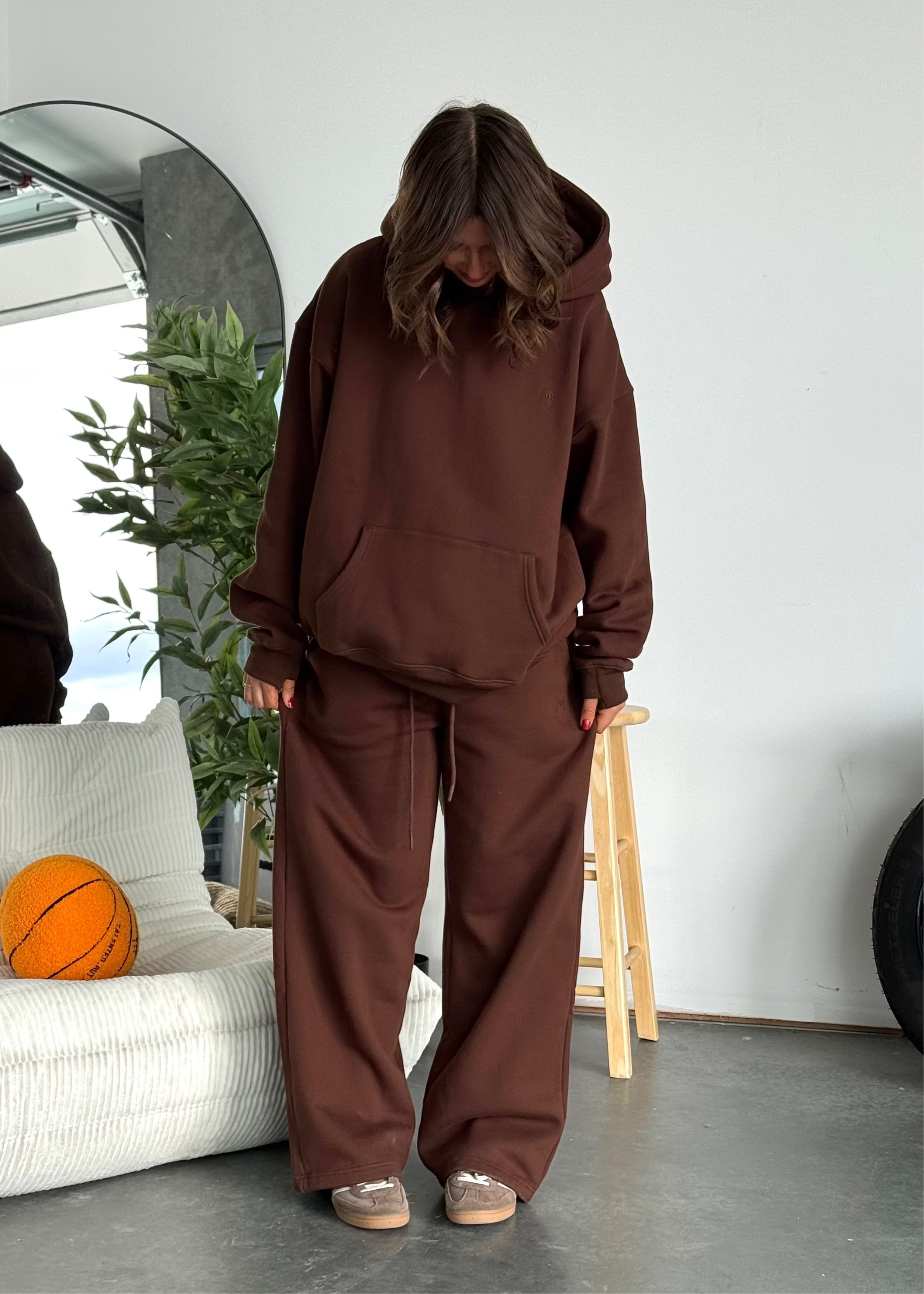Person wearing a brown hoodie and pants standing in a room with a white wall, gray floor, and a plant.