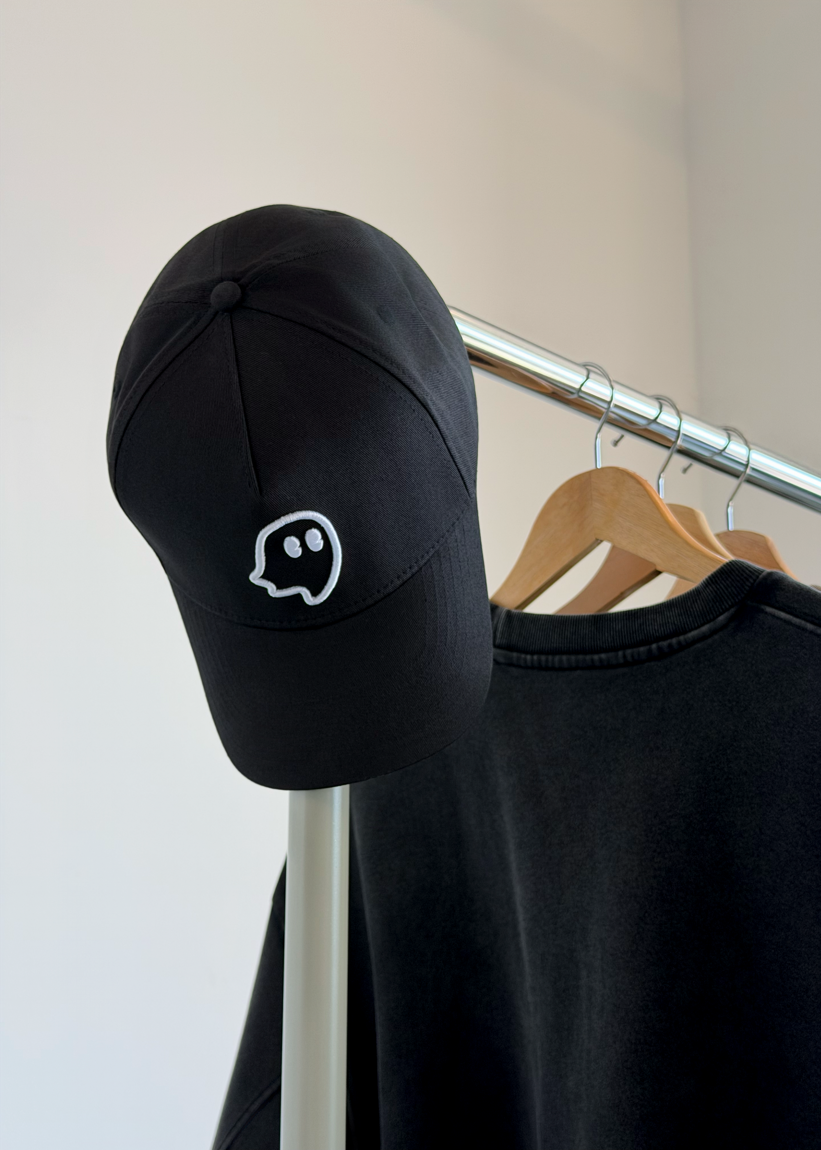 A black baseball cap with a white outlined ghost emoji embroidered on the front, hanging on a clothing rack next to a black sweatshirt.