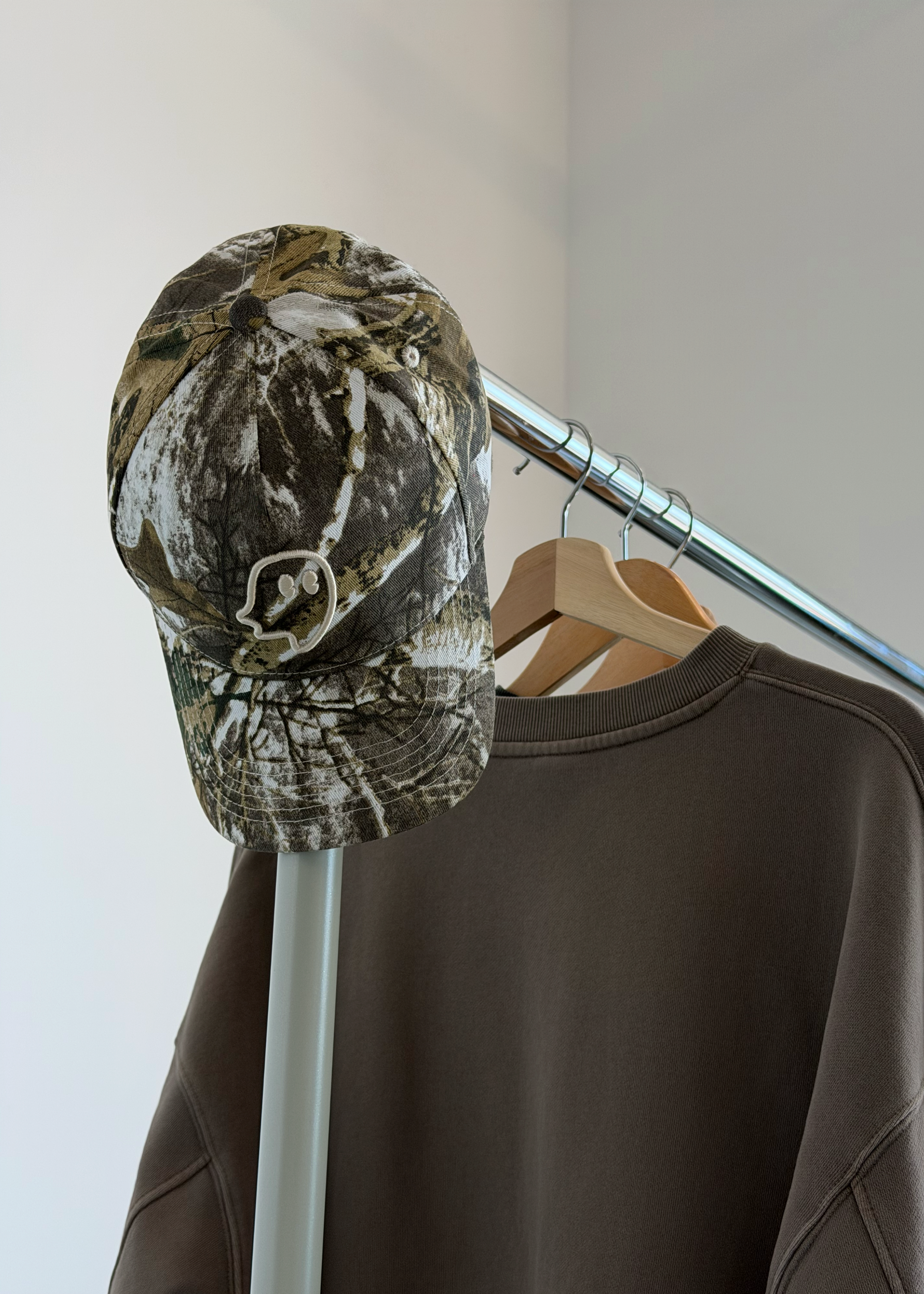 A camouflage baseball cap with a skull logo hanging on a clothing rack next to a brown sweatshirt.